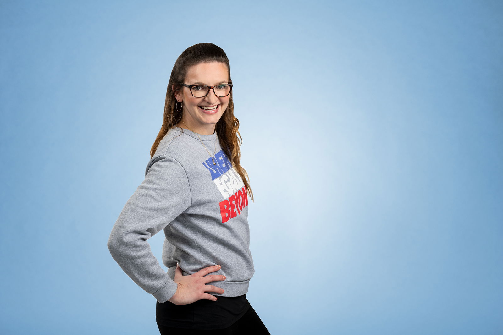 Eine Frau mit langen dunklen Haaren steht seitlich, die Hände in die Hüfte gestemmt, vor einem blauen Hintergrund und lacht über die rechte Schulter in die Kamera. Sie trägt eine Brille. 