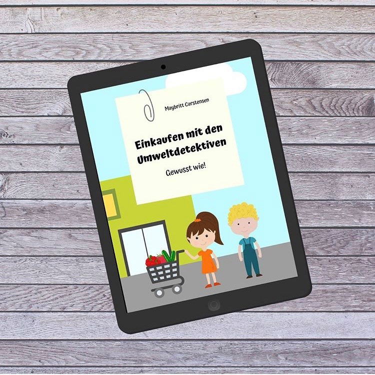 Auf einem Holztisch liegt ein Tablet. Auf dem Tablet sind zwei Menschen mit einem Einkaufswagen. Darüber steht auf einem Zettel Einkaufen mit den Umweltdetektiven.