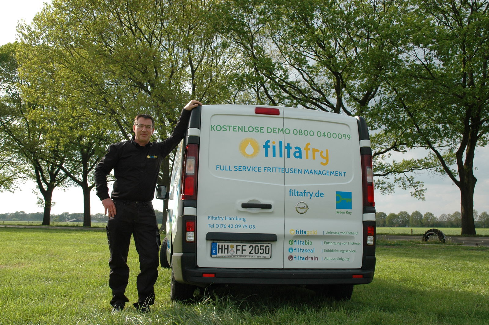 Ein Mann und ein Auto stehen auf einer Rasenfläche. Der Mann lehnt mit seinem linken Arm am Auto und guckt in die Kamera.