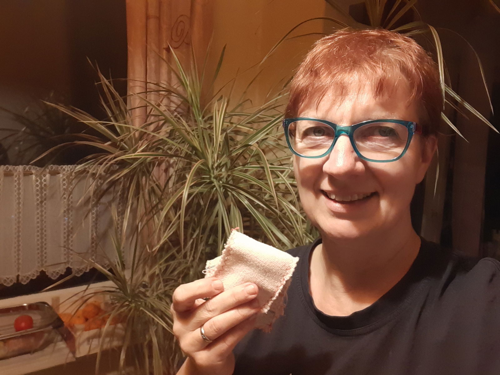 Eine Frau steht vor einer Pflanze. Sie hält zwei Stofflappen in der Hand und lächelt in die Kamera.