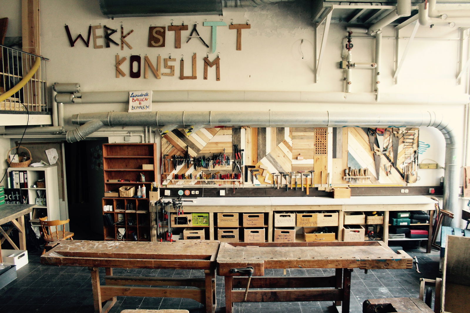 In einem Raum steht ein Holztisch. Am den Wänden hängen viele verschiedene Werkzeuge und Materialien. Darüber hängen selbstgemachte Buchstaben an der Wand, die Werk Statt Konsum darstellen.