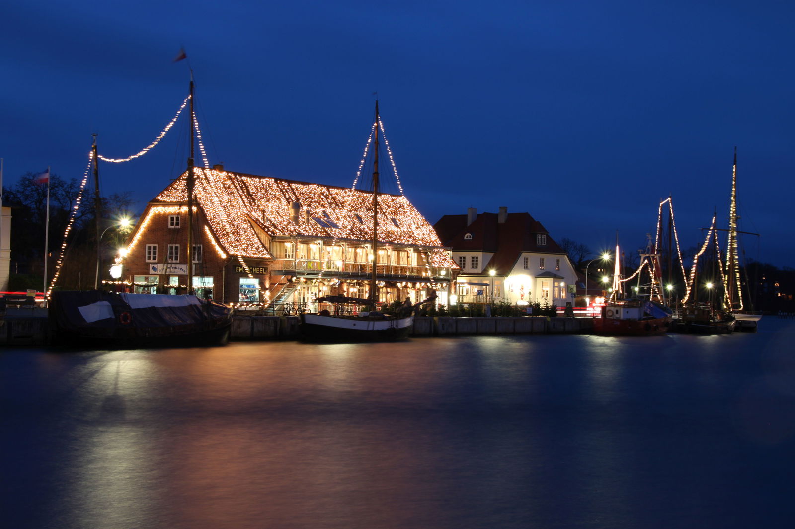 Auf dem Bild ist der Hafen von Neustadt in Holstein am Abend. Im Hafen liegen Segelboote, deren Masten von einer Lichterkette geschmückt werden. Im Hintergrund ist ein Haus, dessen Dach mit einer Lichterkette weihnachtlich geschmückt ist.