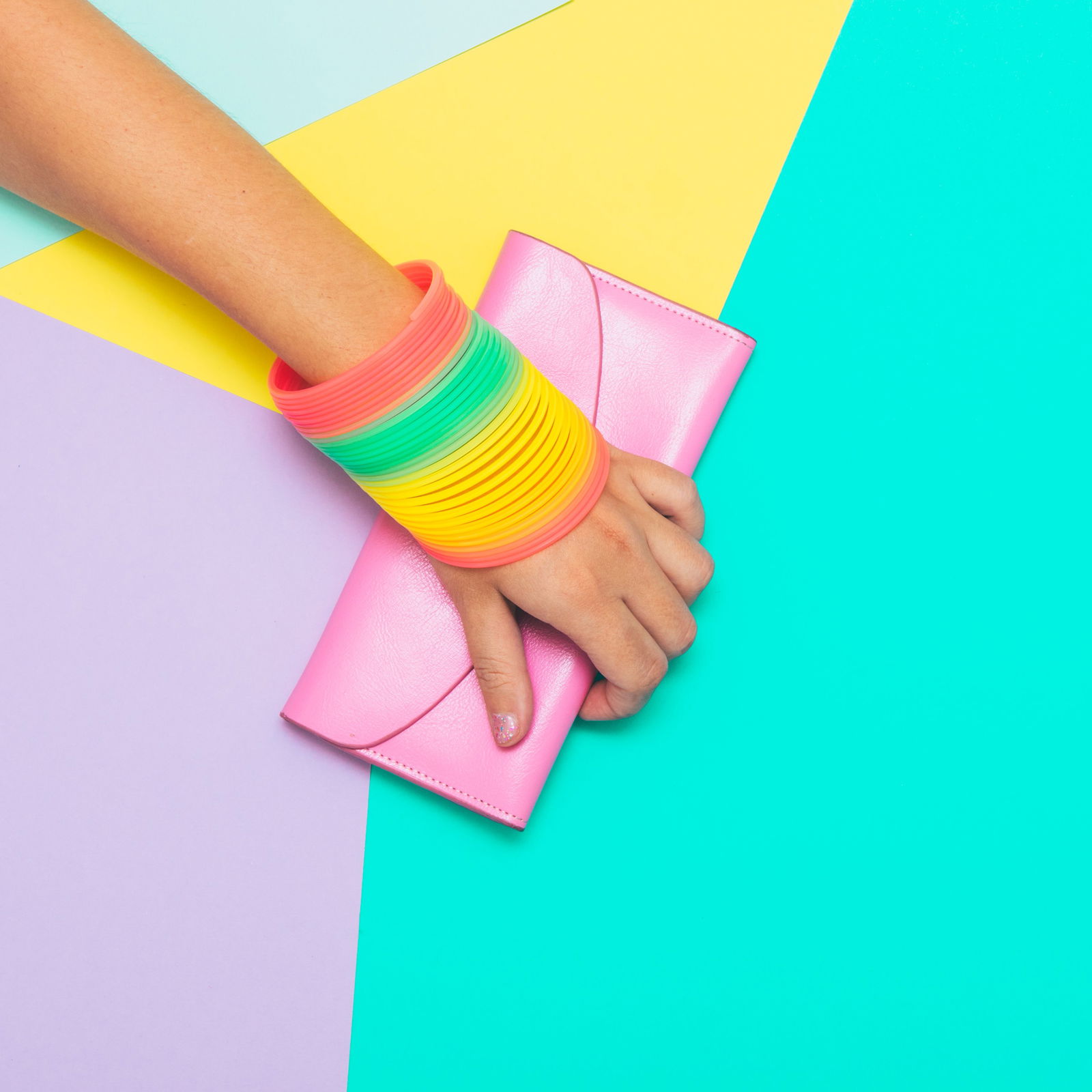 Eine Person streckt von oben links seinen Arm in die Mitte des Bildes. Am Handgelenk trägt sie einen Plastikarmreif in Regenbogenfarben. Die Hand umschließt eine Patellfarbene kleine Handtasche.