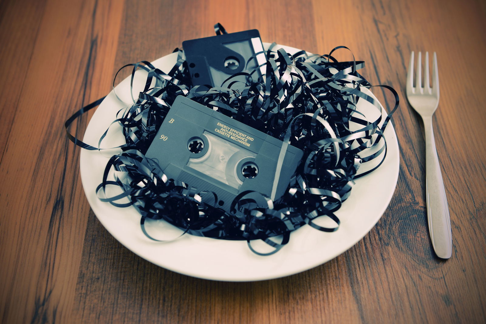 Ein Teller steht auf einem Holztisch liegen zwei Kassetten zwischen Bandsalat. Rechts daneben liegt eine Gabel.