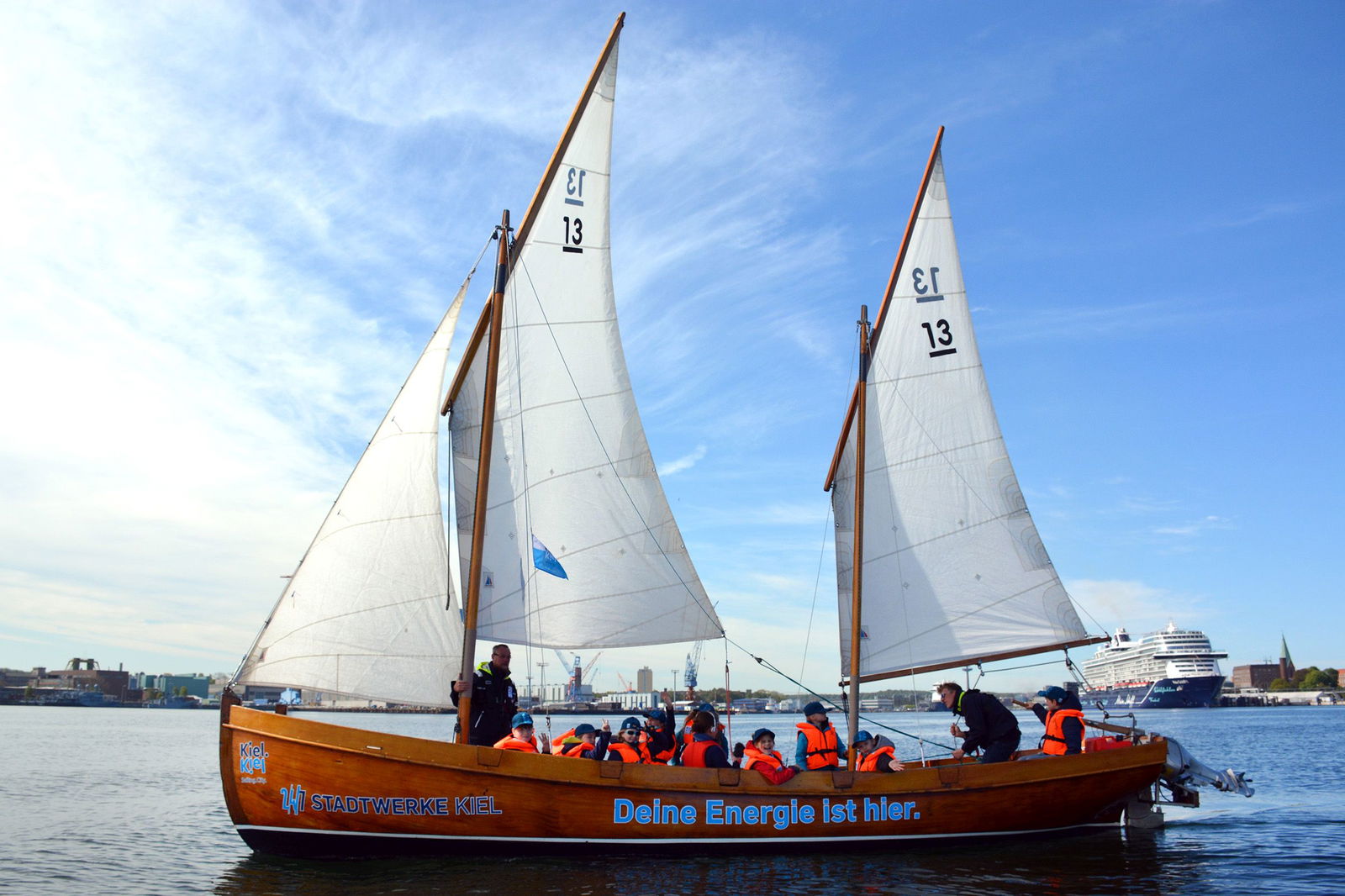 Ein Segelboot fährt mit zwei gehissten Segeln und seiner Besatzung vor einer Küste über das Meer. Rechts im Hintergrund liegt ein Kreuzfahrtschiff vor Anker.