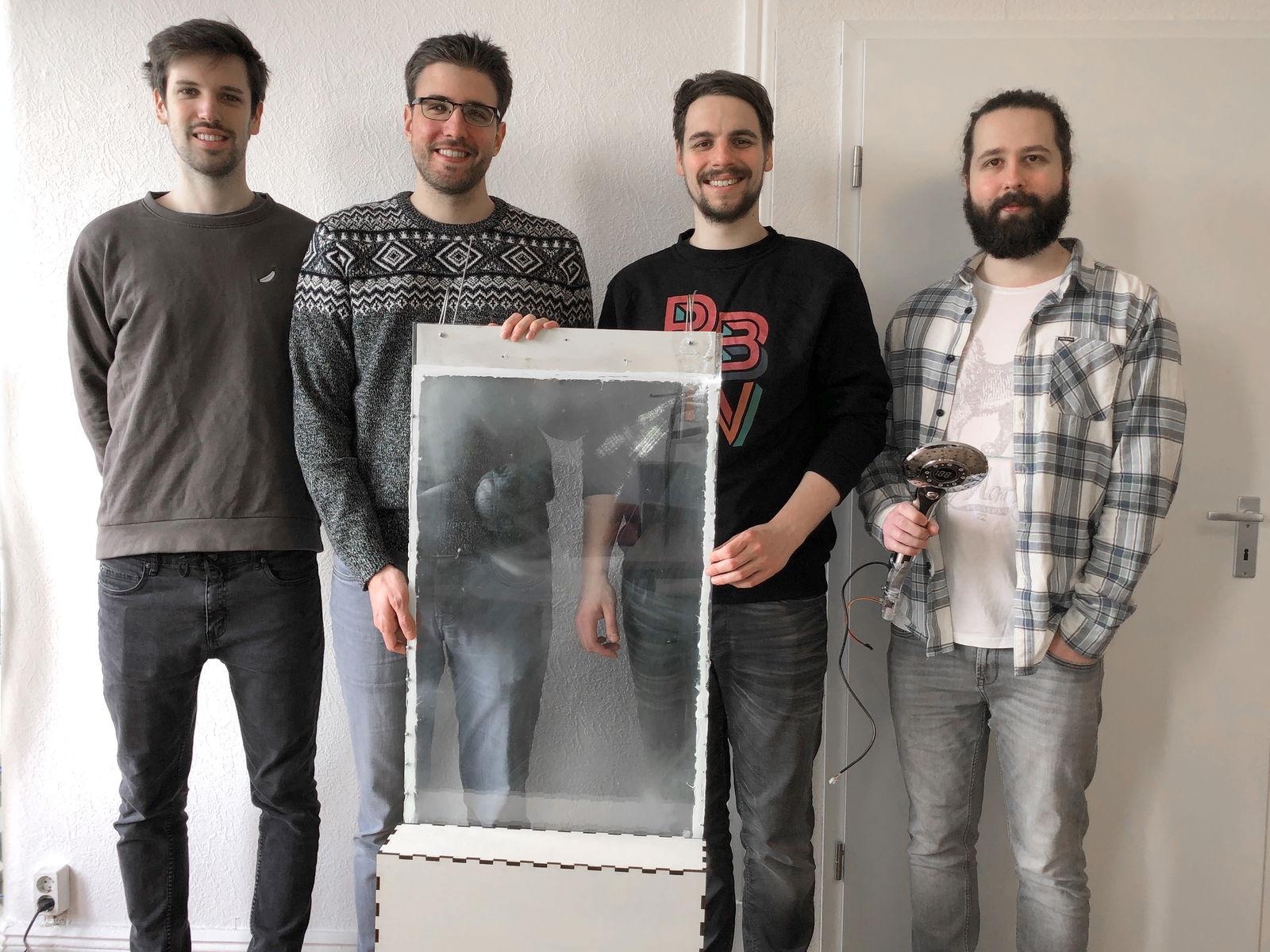 Vier junge Männer stehen nebeneinander vor einer Wand und schauen in die Kamera. Die beiden in der Mitte haben ein selbstgebautes Konstrukt vor sich stehen. Der Mann auf der rechten Seite hat einen Duschkopf in der Hand.