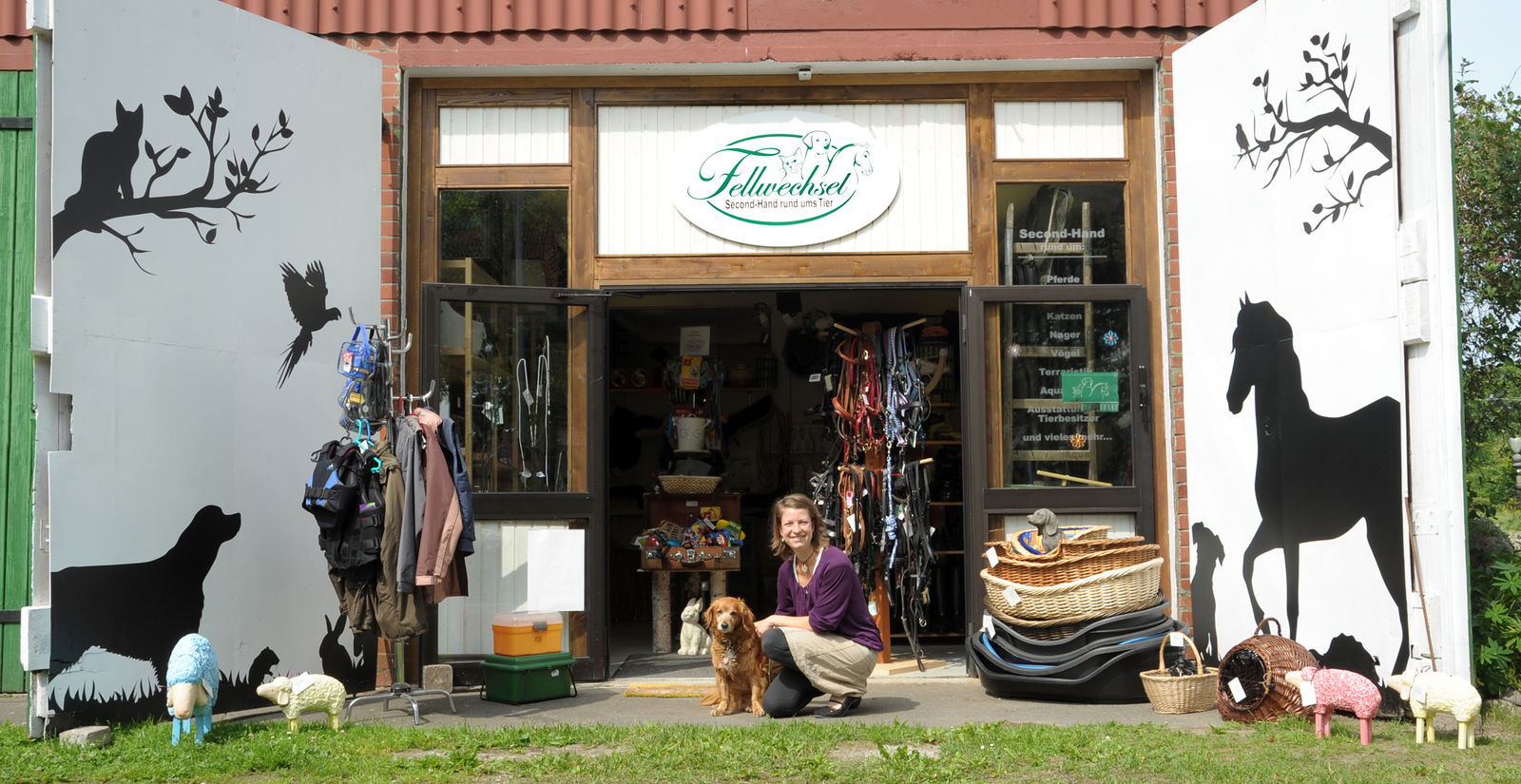 Eine Frau kniet vor einem Haus. Neben ihr sitzt ein Hund und guckt in die Kamera. Neben den beiden stehen sehr viele Körbchen und im Haus ist ein Laden, in dem viele Hundeleinen und weitere Dinge für Tiere sind.