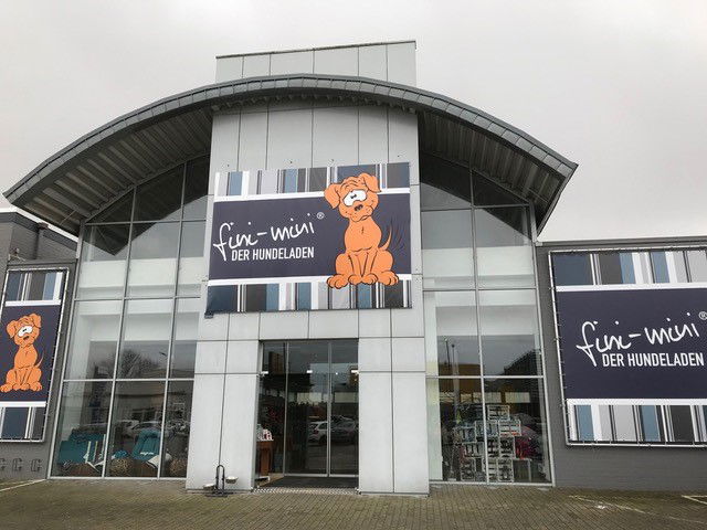Über dem Eingang eines Gebäudes ist ein großes Schild angebracht. Auf dem Schild ist ein Hund abgebildet. Links neben dem Hund steht "fini-mini Der Hundeladen".