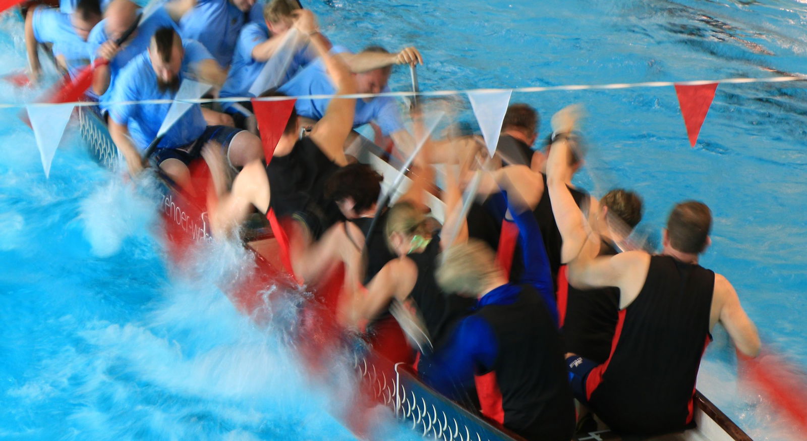 Zwei Mannschaften sitzen in einem Drachenboot und rudern in einem Indoorpool gegeneinander.