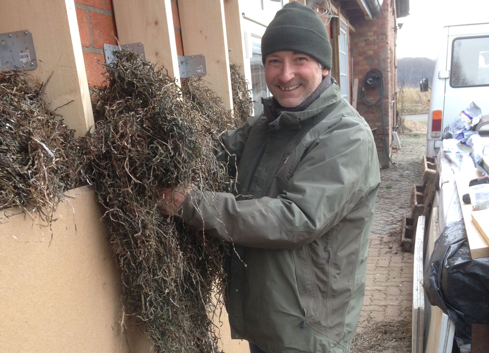 Ein Mann steht an einem Haus und hält Seegras in den Händen. Er lächelt in die Kamera. 