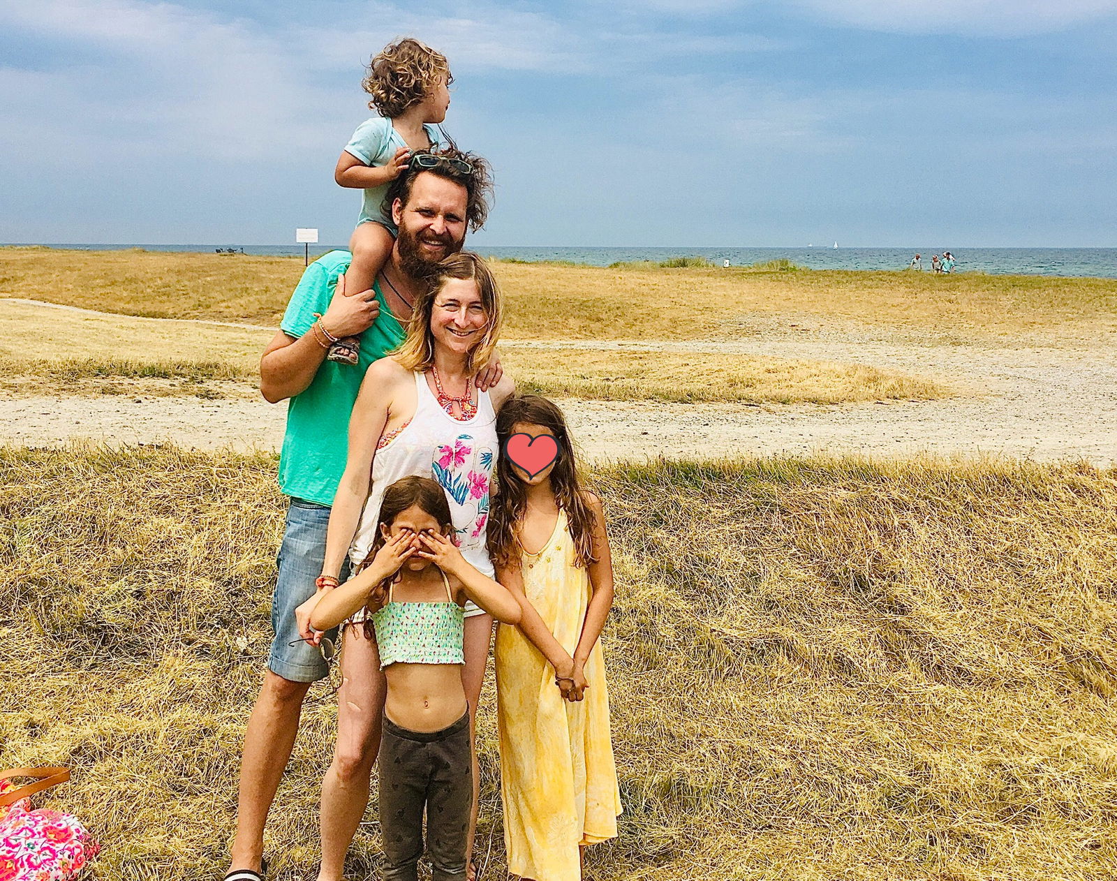 Ein Mann und eine Frau stehen an einem Strand. Auf den Schultern des Mannes sitzt ein kleines Kind. vor der Frau stehen zwei kleine Mädchen.