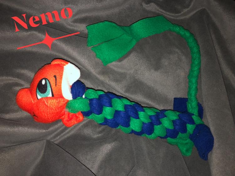 Auf einem grauen Hintergrund liegt ein selbstgebastelter Fisch. Dieser ist aus einer grünen und blauen Pferdebandage zusammengeknüpft und hat einen orange-weißen Kopf.