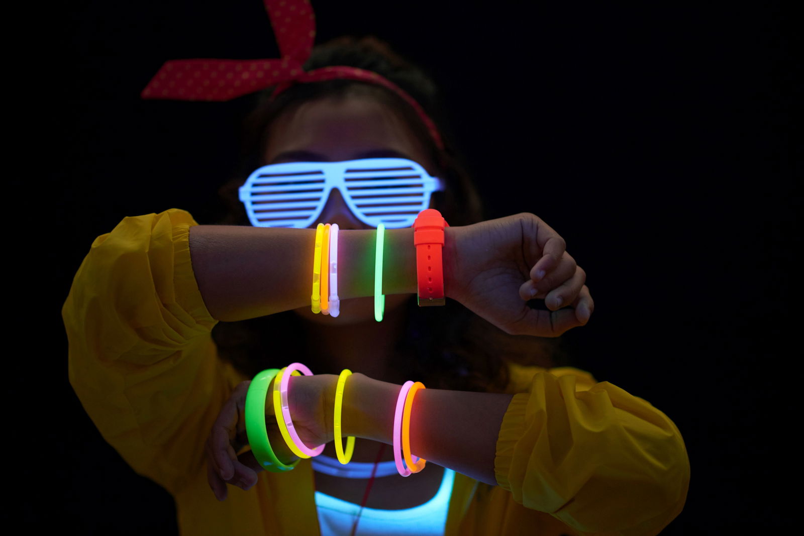 Eine Person hält sich im Dunkeln beide Arme vor das Gesicht. An den Handgelenken trägt sie fluoreszierende Armbänder, die bunt leuchten. Zusätzlich trägt sie eine Sonnebrille, die ebenfalls leuchtet.