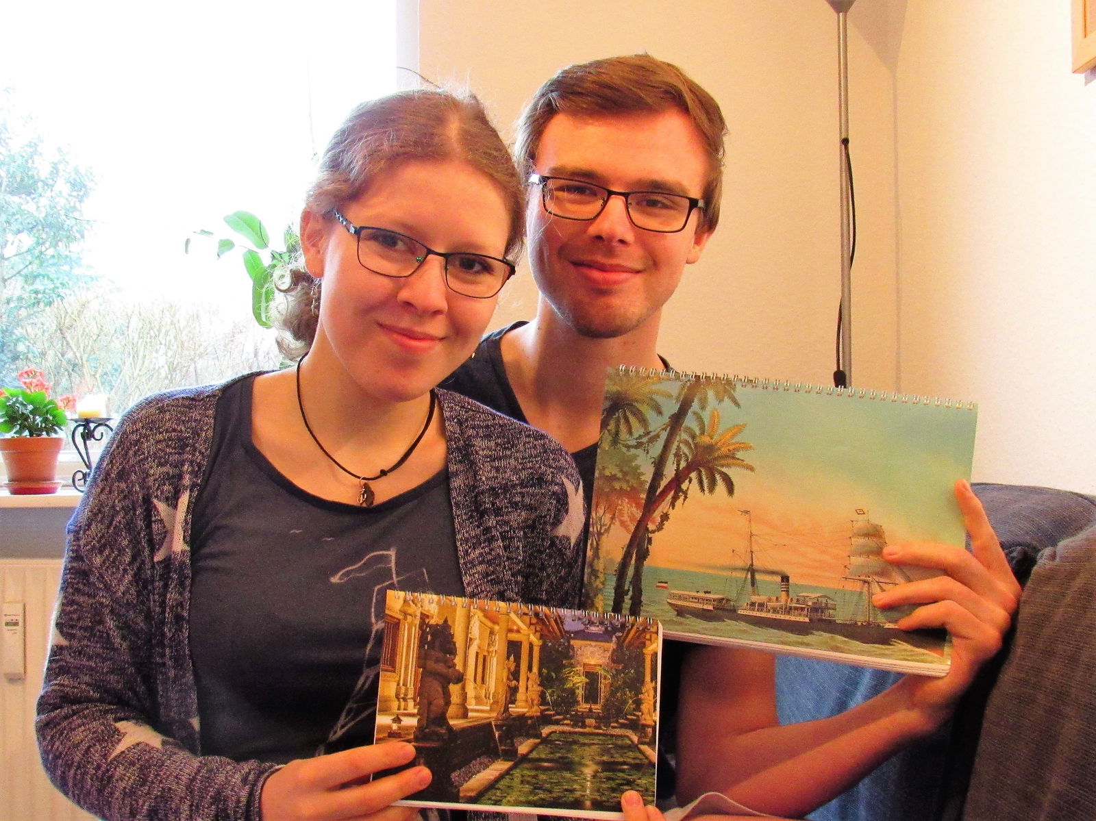 Zwei Personen sitzen auf einem Sofa und halten jeweils einen Notizblock in ihrer Hand.