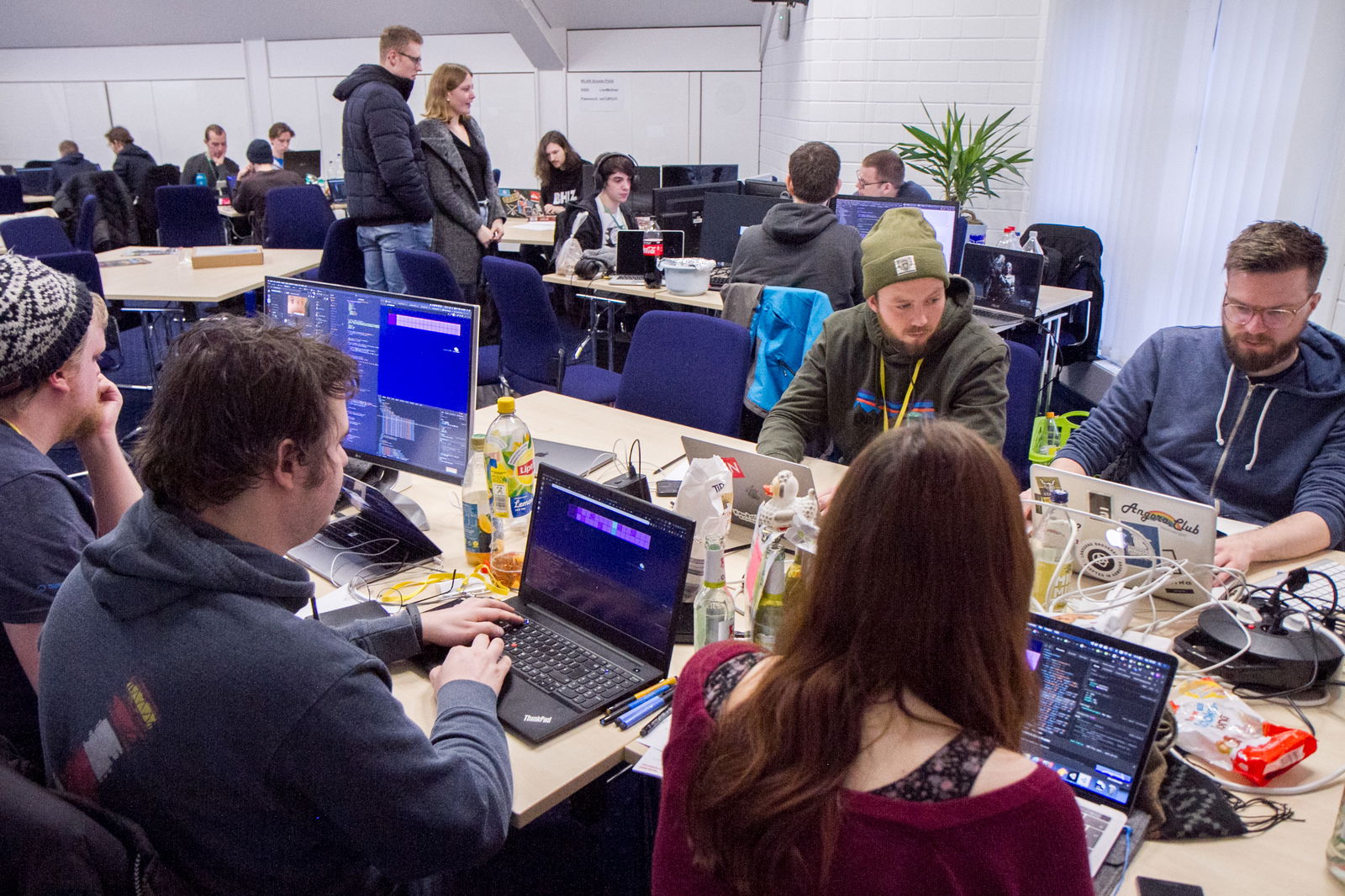 An einem Tisch sitzen fünf Personen, die an Laptops arbeiten. Im Hintergrund sind weitere Tische, an denen Menschen sitzen. An einem Tisch stehen zwei Personen und reden mit den anderen am Tisch.