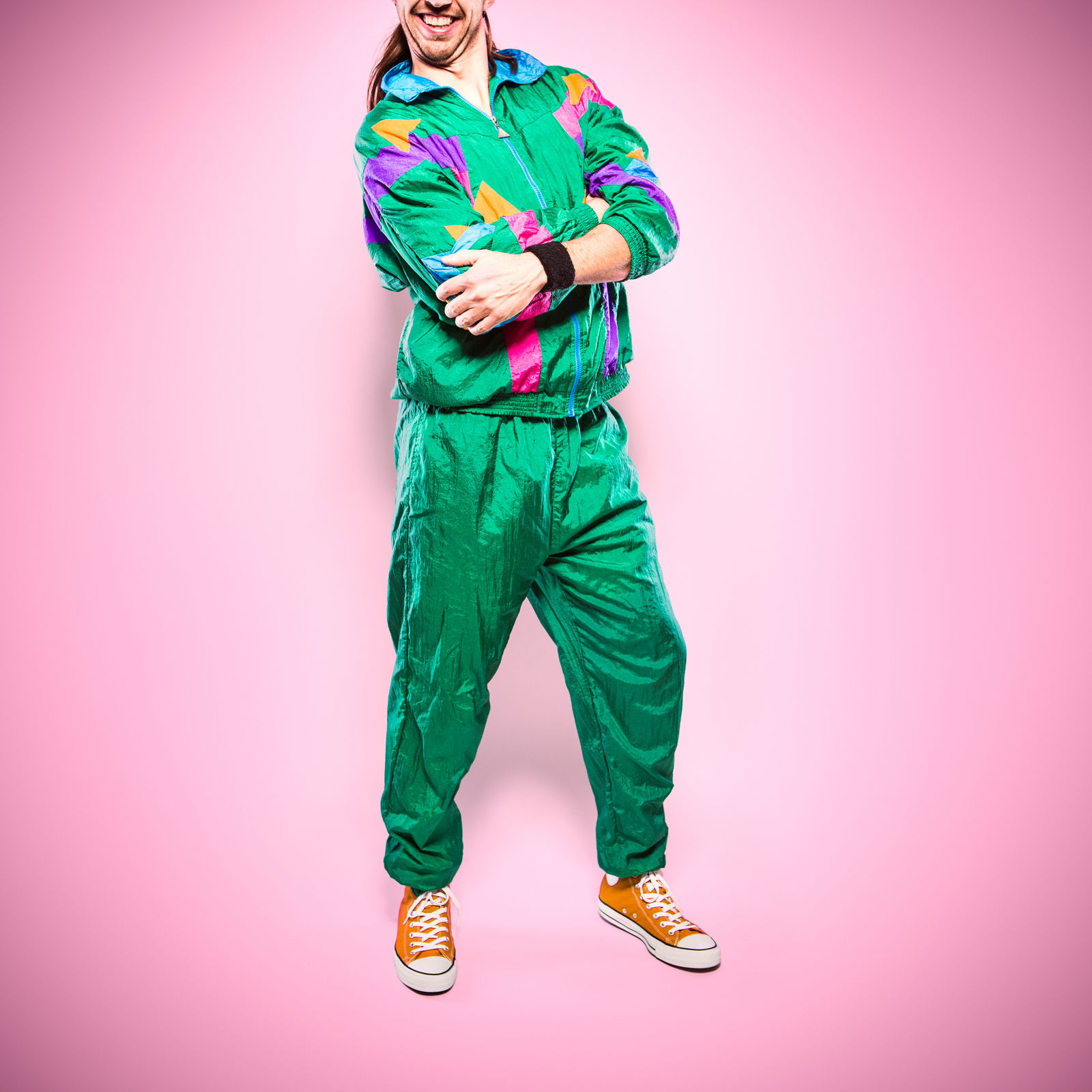 Eine Person ist mit einem bunten, zweiteiligen Nylontrainigsanzug bekleidet und steht mit verschränkten Armen der Kamera zugewandt. 