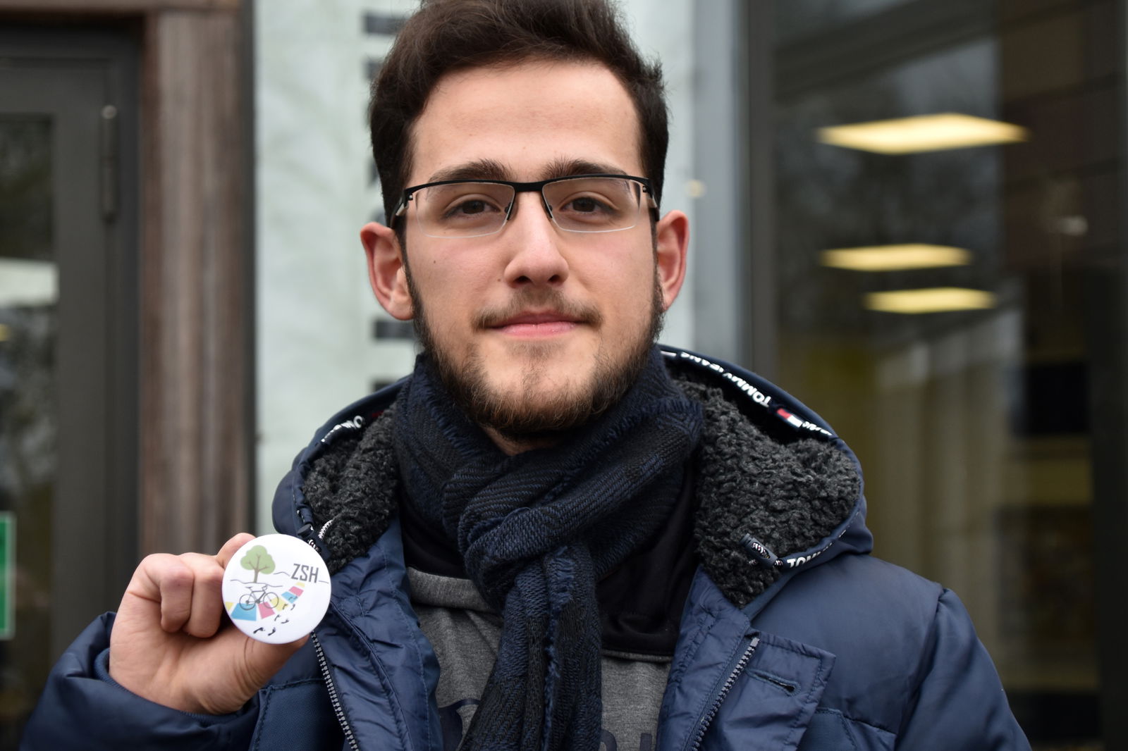 Ein junger Mann mit Brille und Bart guckt in die Kamera und hält mit der rechten Hand einen kleinen Button ins Bild.