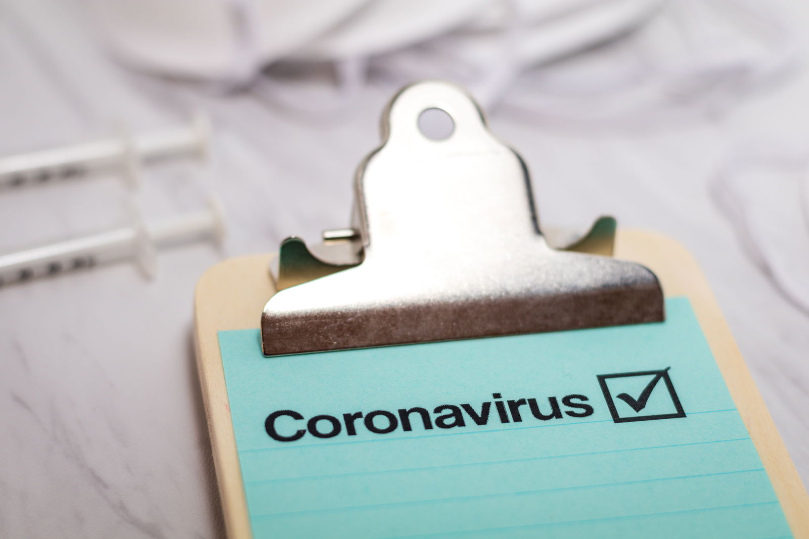 Auf einem Klemmbrett klemmt ein blaues Papier mit dem Titel "Coronavirus". Der Untergrund ist grau und im Hintergrund liegen zwei Spritzen.