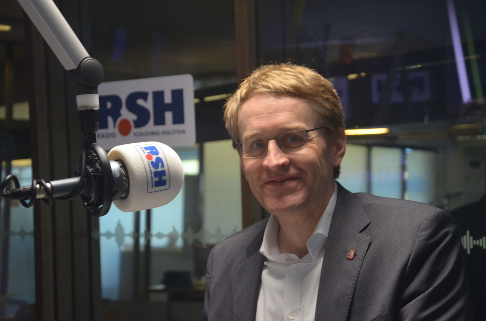 Ein Mann in mittlerem Alter, ist mit Anzug und Hemd bekleidet. Er sitzt in einem Studio vor einem Mikrofon und schaut in die Kamera.