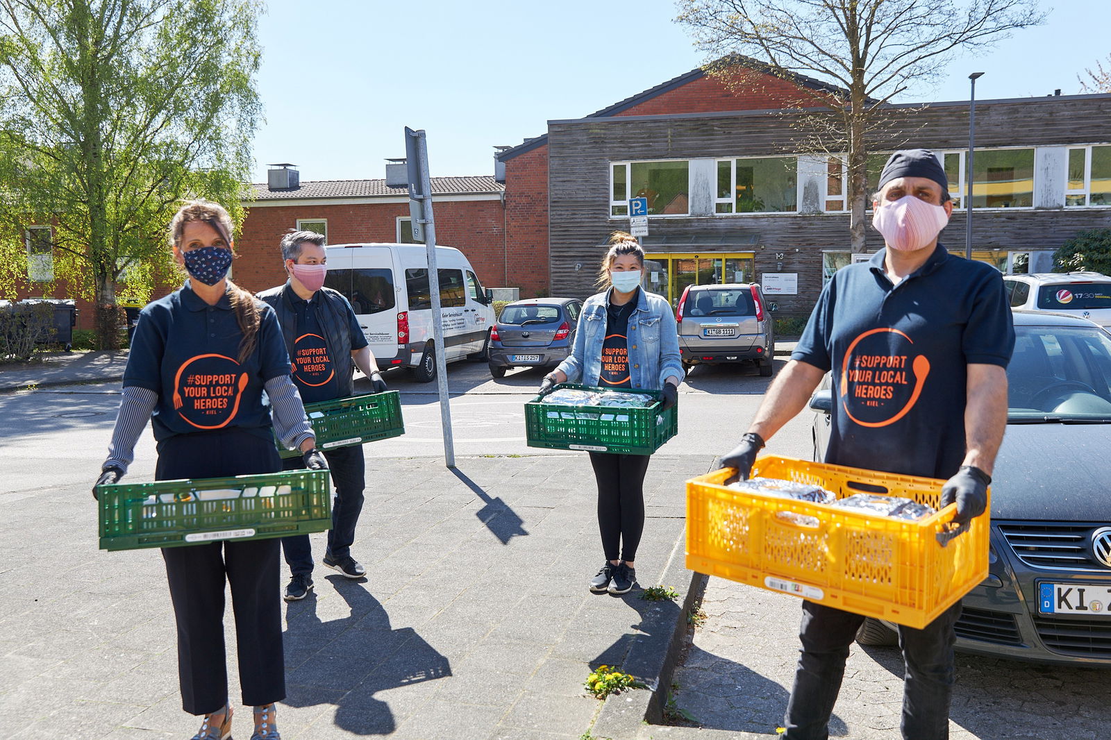 Zwei Männer und Zwei Frauen stehen auf einem Parkplatz. Sie halten grüne und gelbe Kisten voll Lebensmittel vor ihrem Körper. Alle tragen ein dunkelblaues T-Shirt mit der Aufschrift "Support your local heroes" sowie einen Mund-und-Nasen-Schutz.
