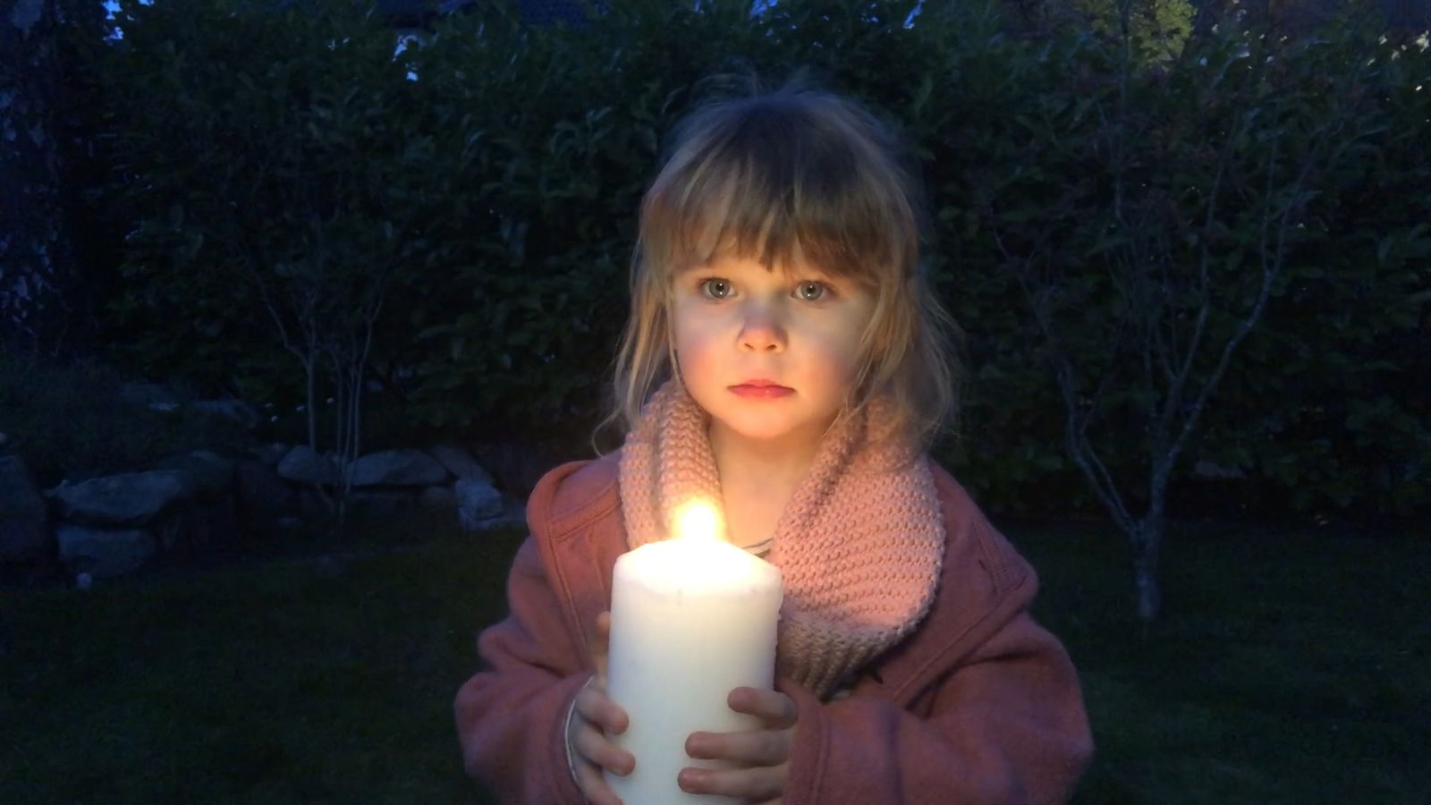 Ein kleines Mädchen steht in einem dunklen Garten, schaut in die Kamera und hält mit beiden Händen eine brennende Kerze vor sich.