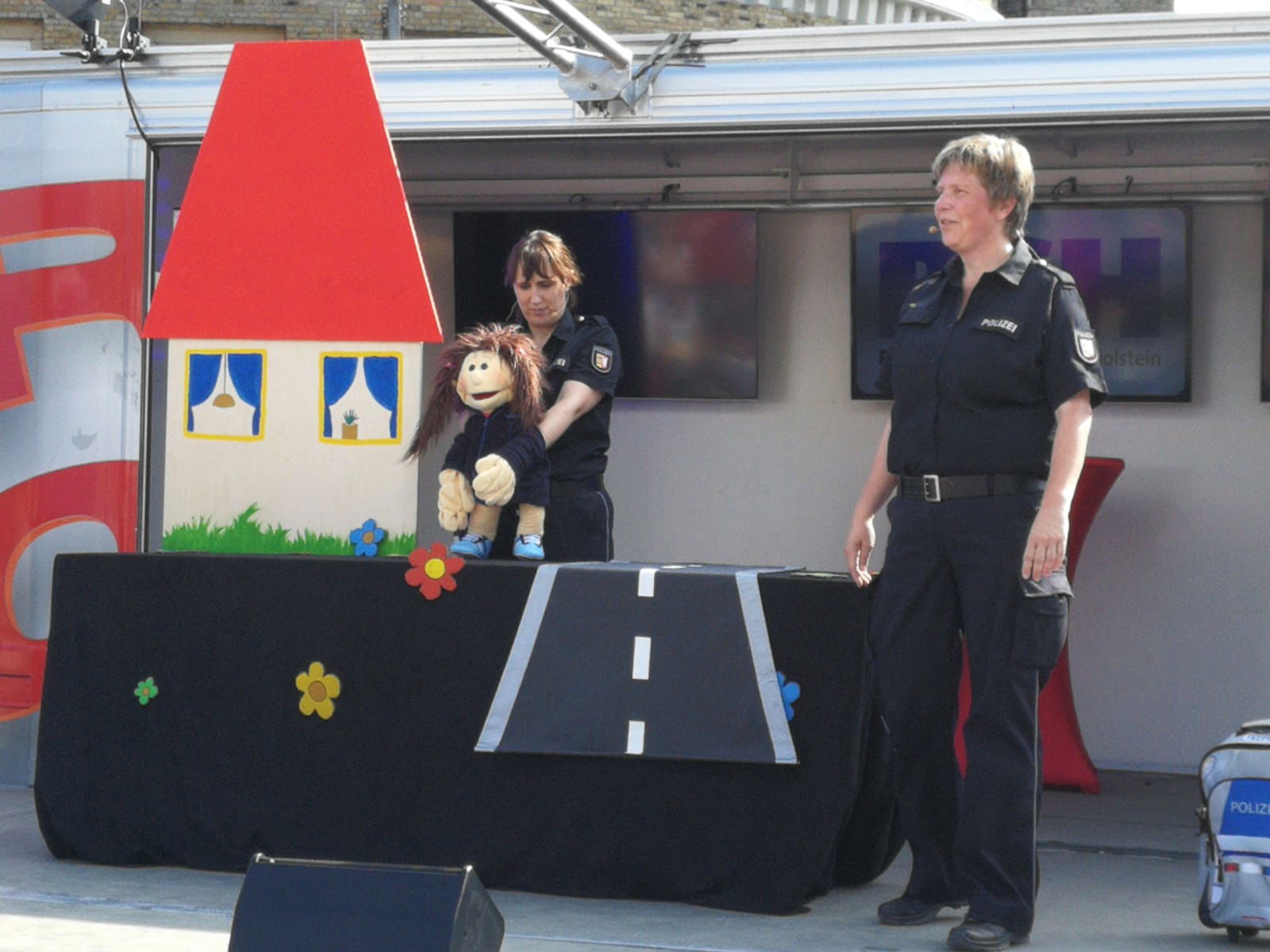 Auf einer Bühne steht eine Puppenspielbühne hinter der eine Frau in Polizeiuniform ein Theaterstück mit einer großen Handpuppe spielt. Rechts daneben steht eine Frau in Polizeiuniform die in ein Mikro an einem Headset spricht.