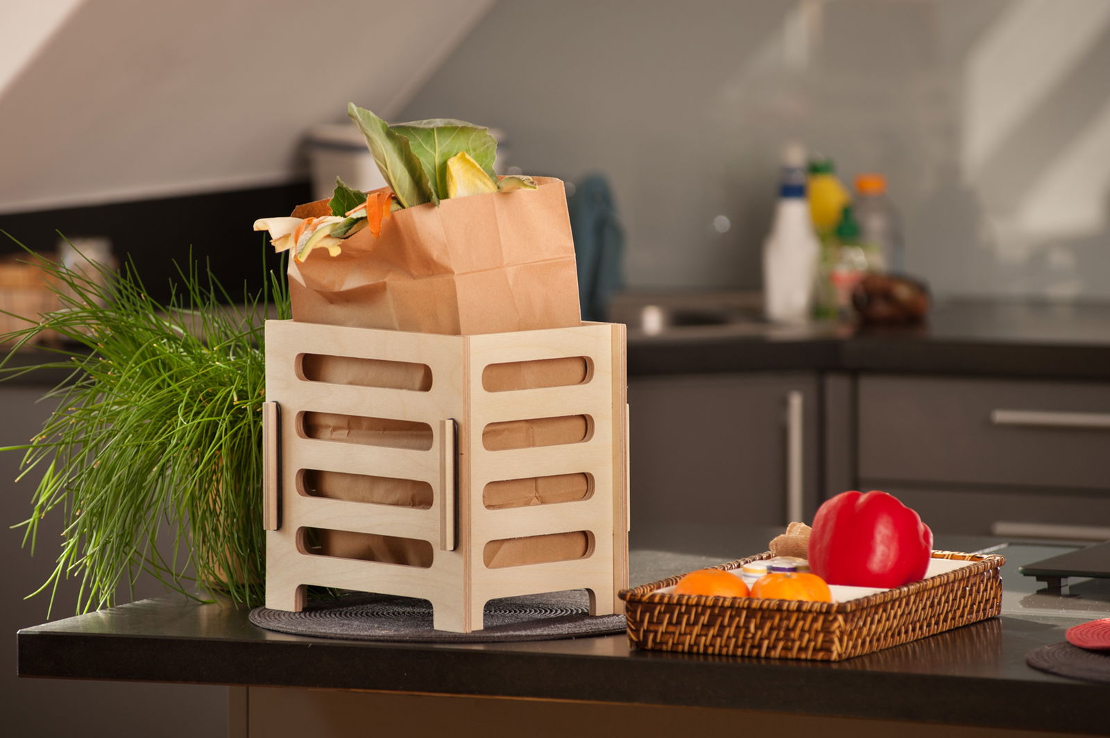 Auf einer Küchentheke steht ein viereckiges Gestell aus Holz. Darin befindet sich eine Biomüll-Tüte mit verschiedenen Bio-Abfällen drin. 