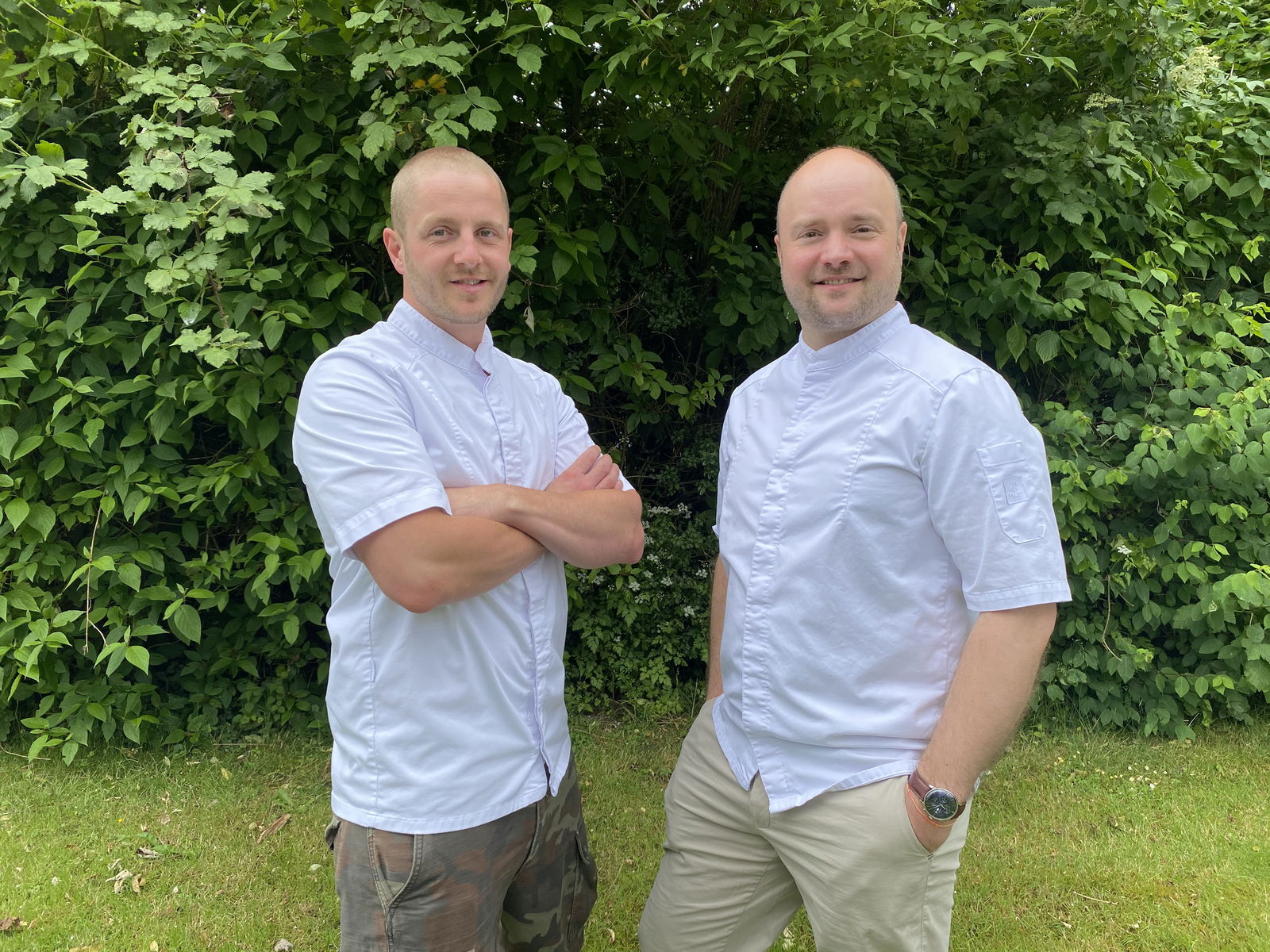Zwei Männer stehen nebeneinander in einem Garten vor einer grünen Hecke.