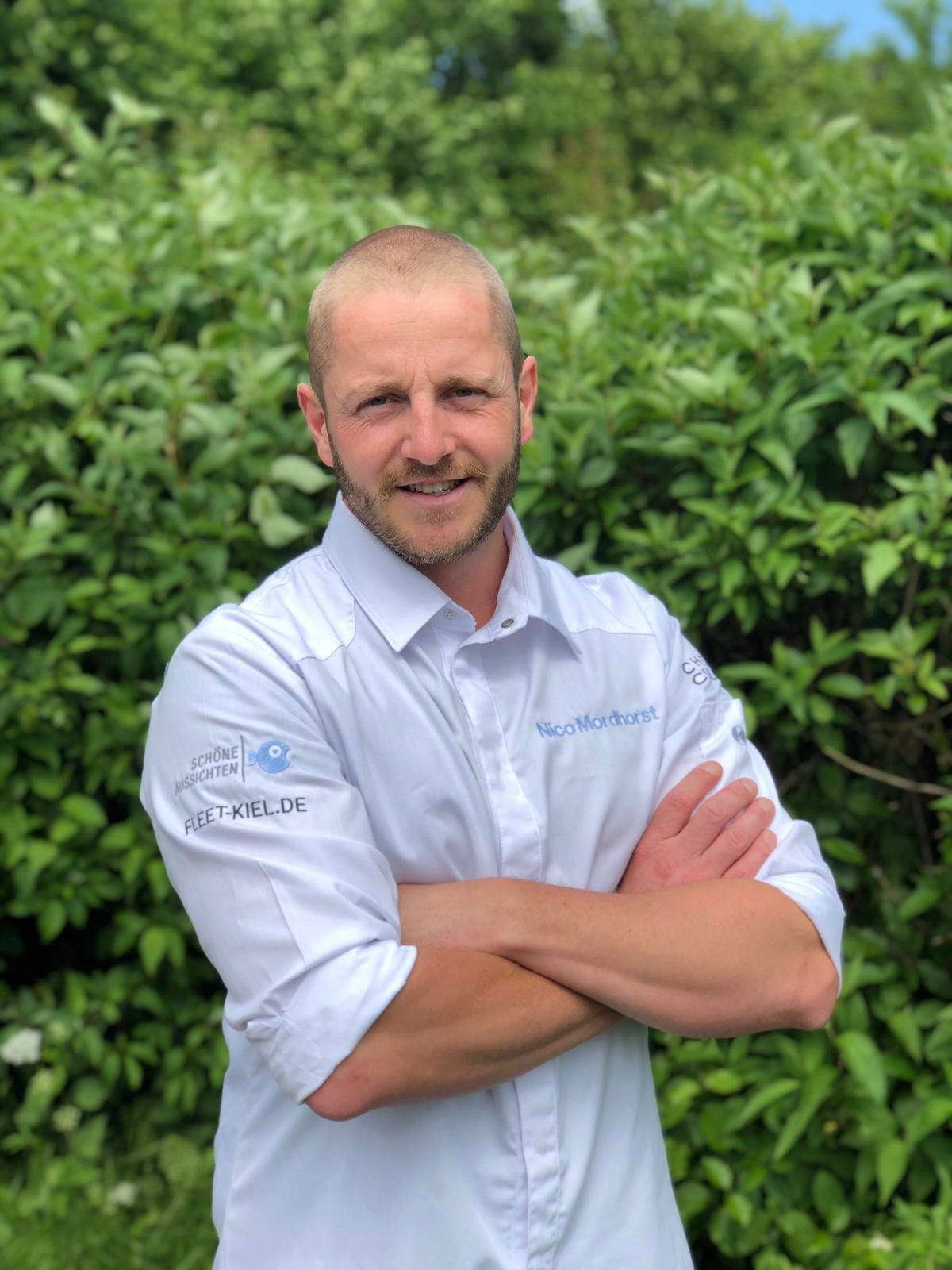 Vor grünem Vordergrund steht ein kurzhaariger Mann mit verschränkten Armen. Auf seiner Kochjacke steht "Nico Morshorst".