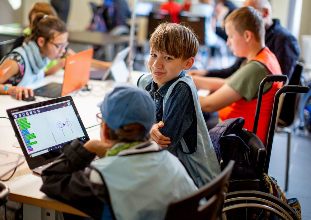Ein Junge im Rollstuhl schaut freundlich in die Kamera. Um ihn herum sitzen andere Kinder, die auf Laptops schauen.