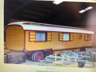 Ein alter Zirkuswagen steht in einer Halle. Der Zirkuswagen ist mit gelben Holzplanken von außen verziert, hat Fenster mit Fensterläden und ein weißes Dach.