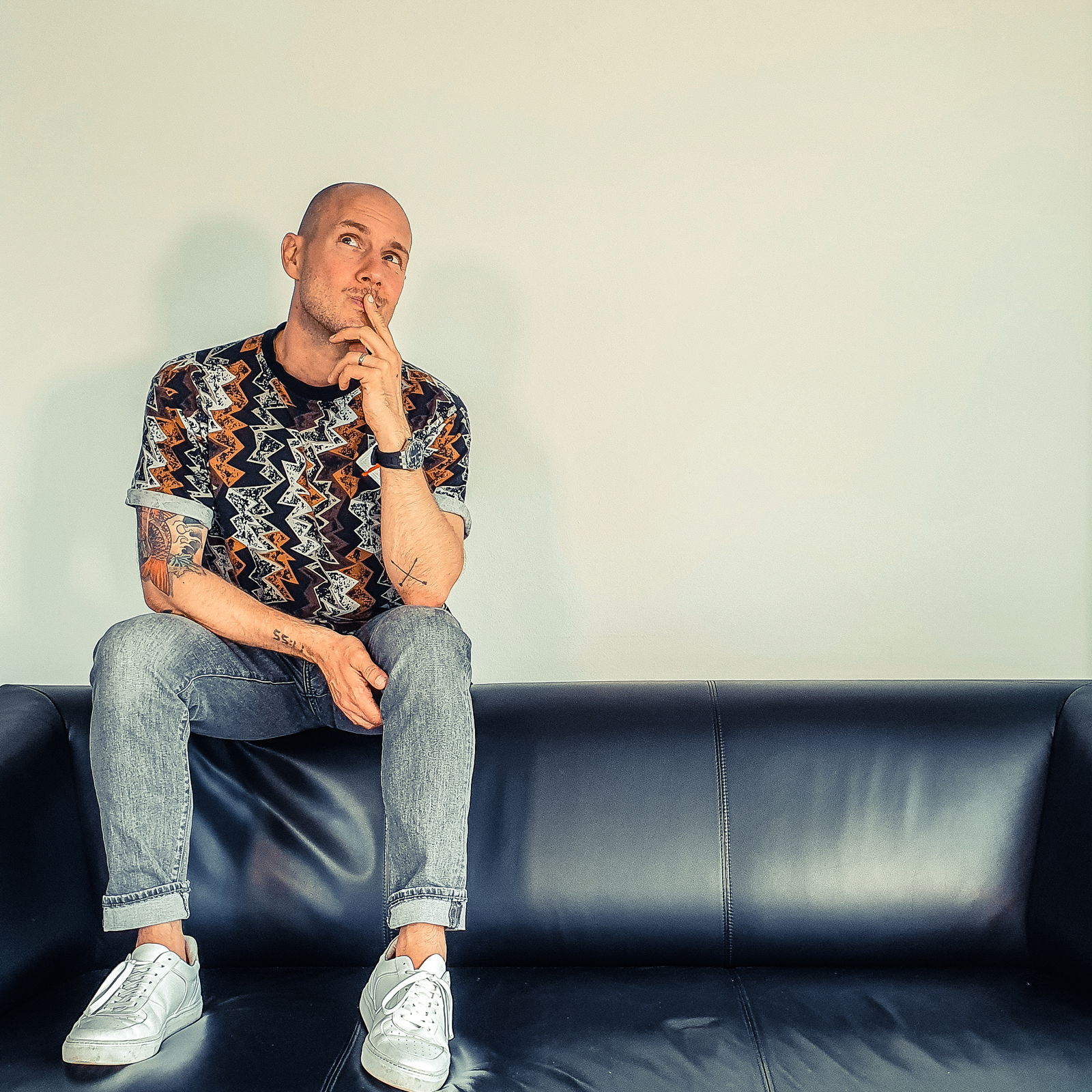 Oli.P sitzt auf der Lehne eines schwarzen Sofas und hat nachdenklich eine Hand am Kinn.