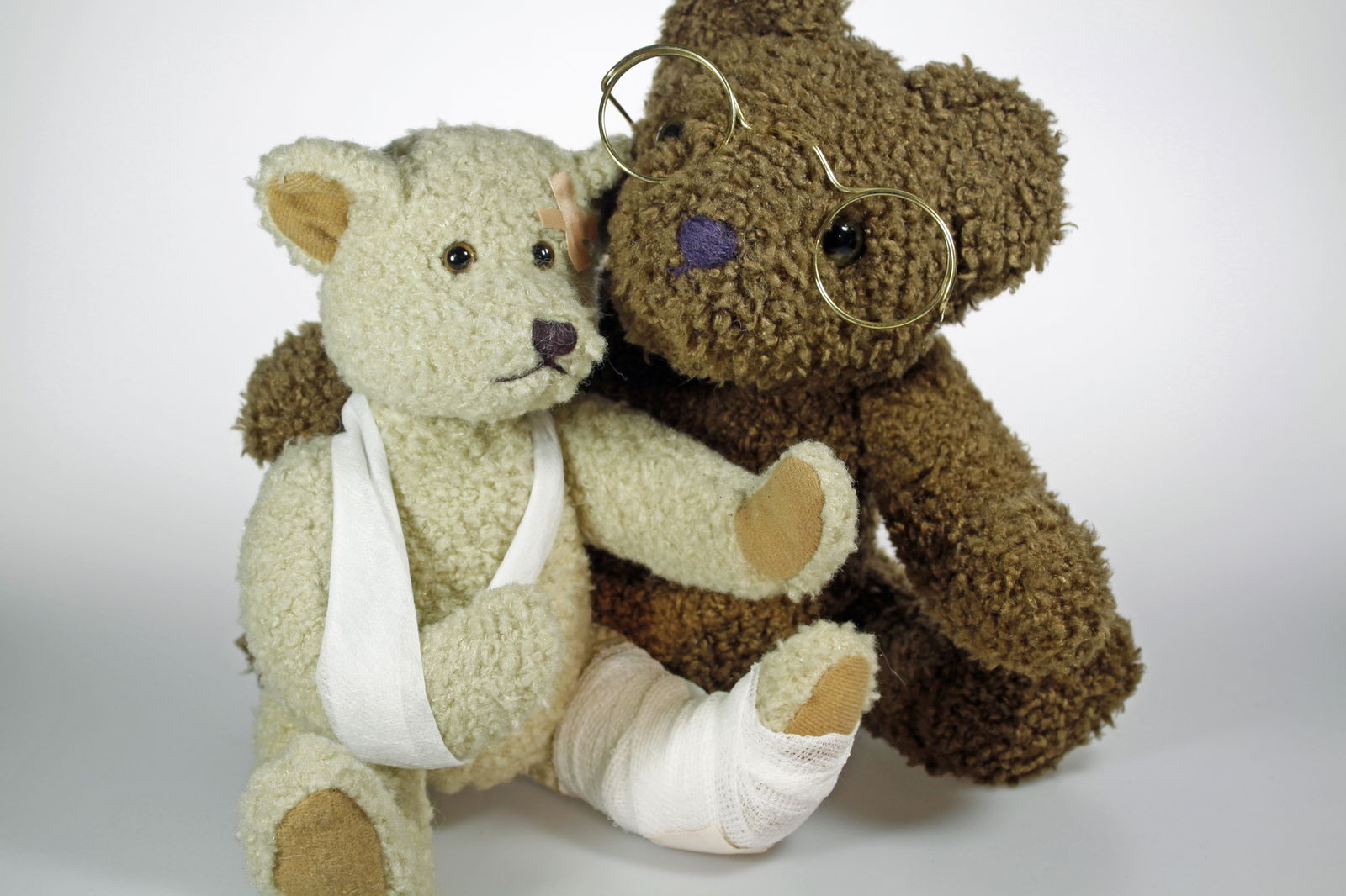 Ein brauner Teddybär mit Brille nimmt einen beigen Teddybär mit Verband in den Arm. Beide sitzen auf dem Boden.
