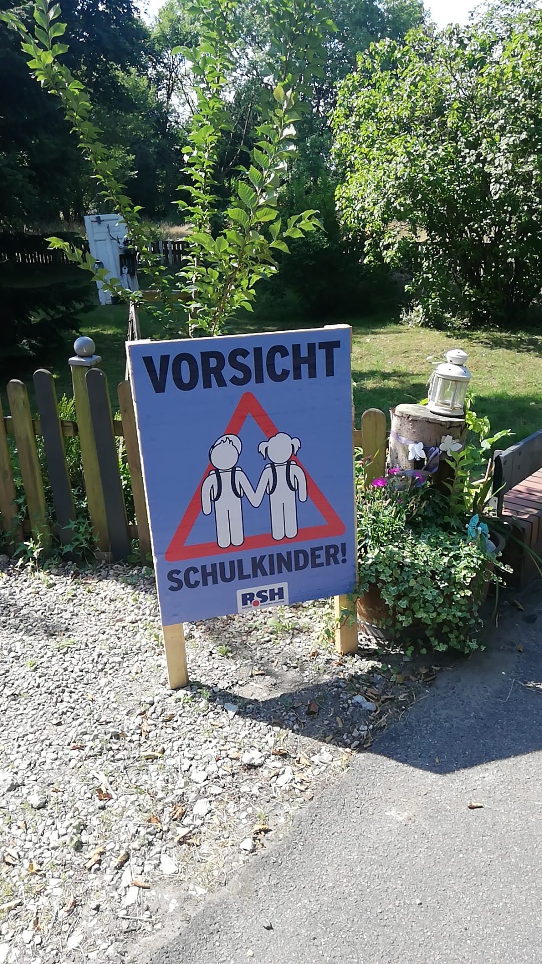 Ein Plakat mit der Aufschrift "Vorsicht Schulkinder" steht auf Schotter vor einem Garten.
