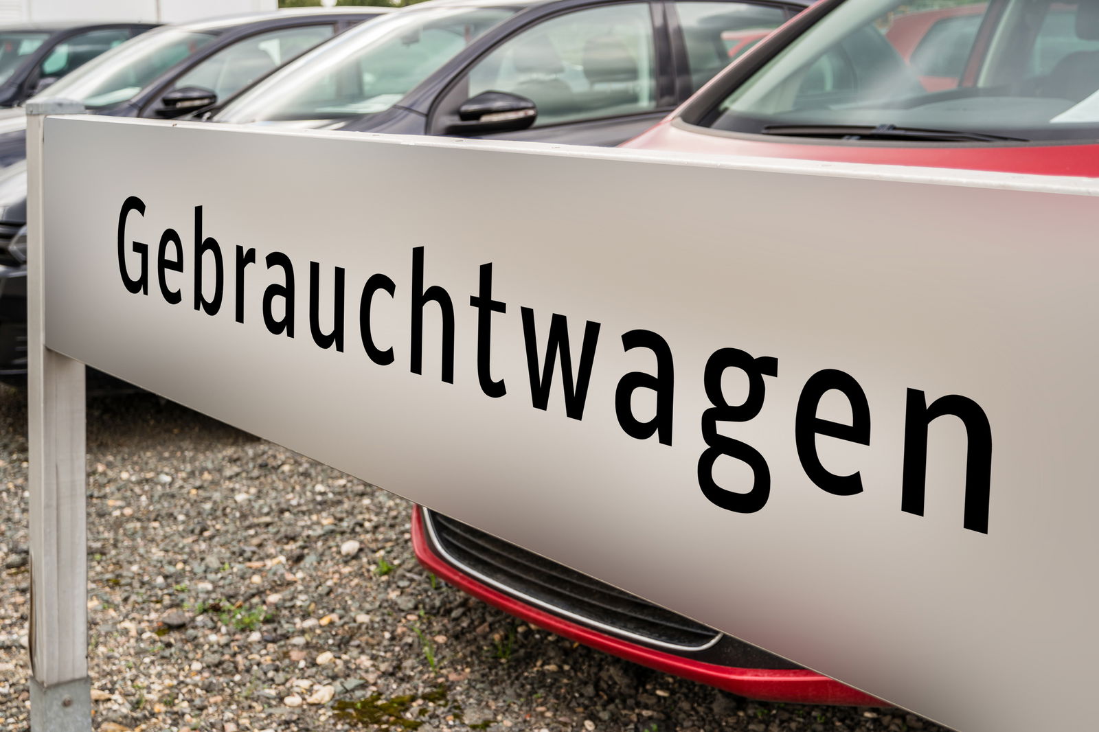 Auf einem weißen Schild steht das Wort Gebrauchtwagen. Dahinter stehen Autos.