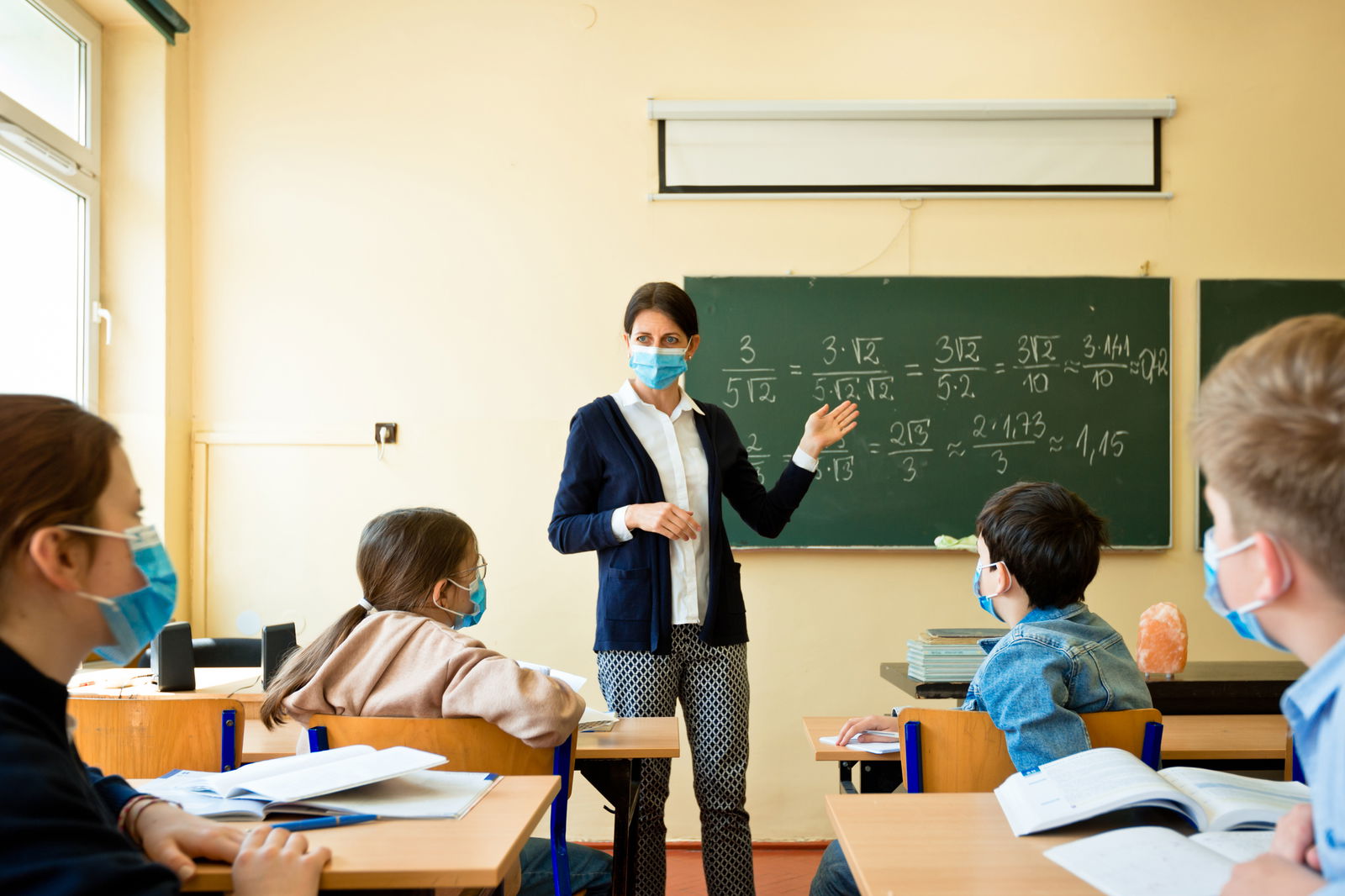 Eine Lehrerin steht vor einer beschrifteten Tafel und schaut die vier Schüler an, die an ihren Tischen sitzen. Alle tragen Atem-Masken.