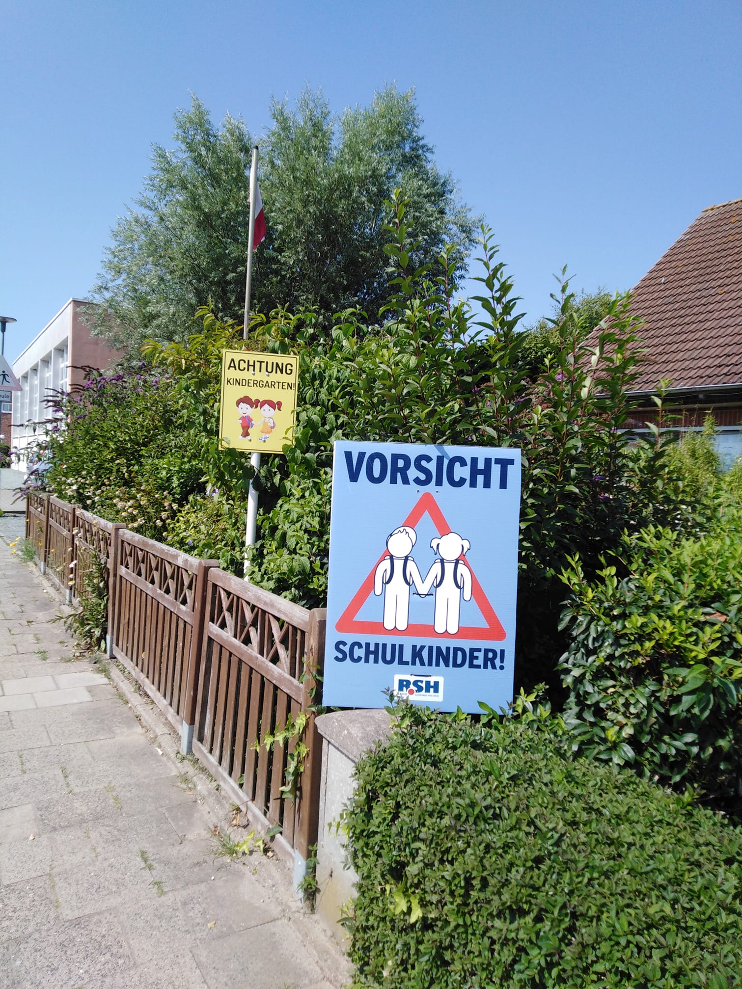 Ein Plakat mit der Aufschrift "Vorsicht Schulkinder" steht auf einem Gartenzaun an einer Straße.