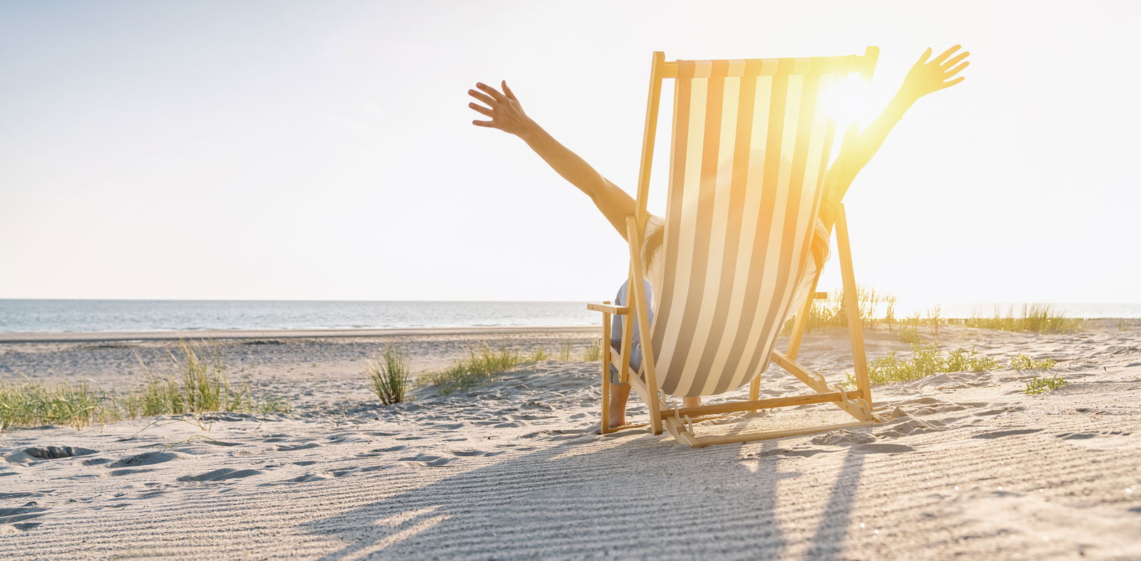 Ein Mensch sitzt auf einem Liegestuhl am Strand und streckt die Arme nach oben. Dabei blickt er auf das Meer und genießt die helle Sonne am blauen Himmel