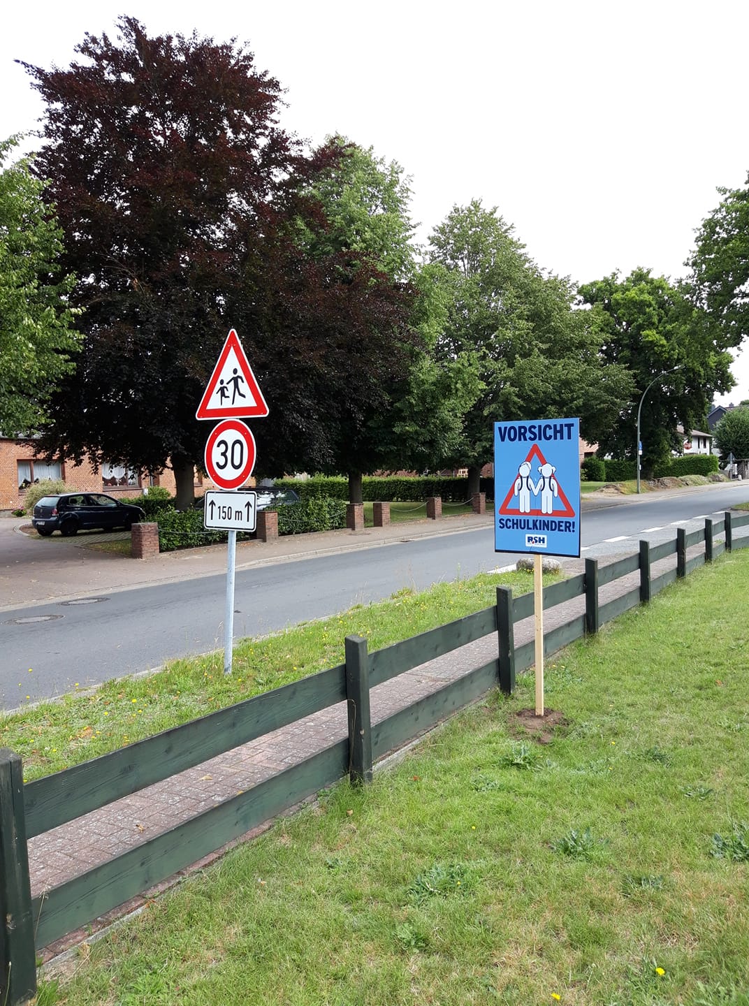 Ein Plakat mit der Aufschrift "Vorsicht Schulkinder" steht an einem Gartenzaun an einer Straße.