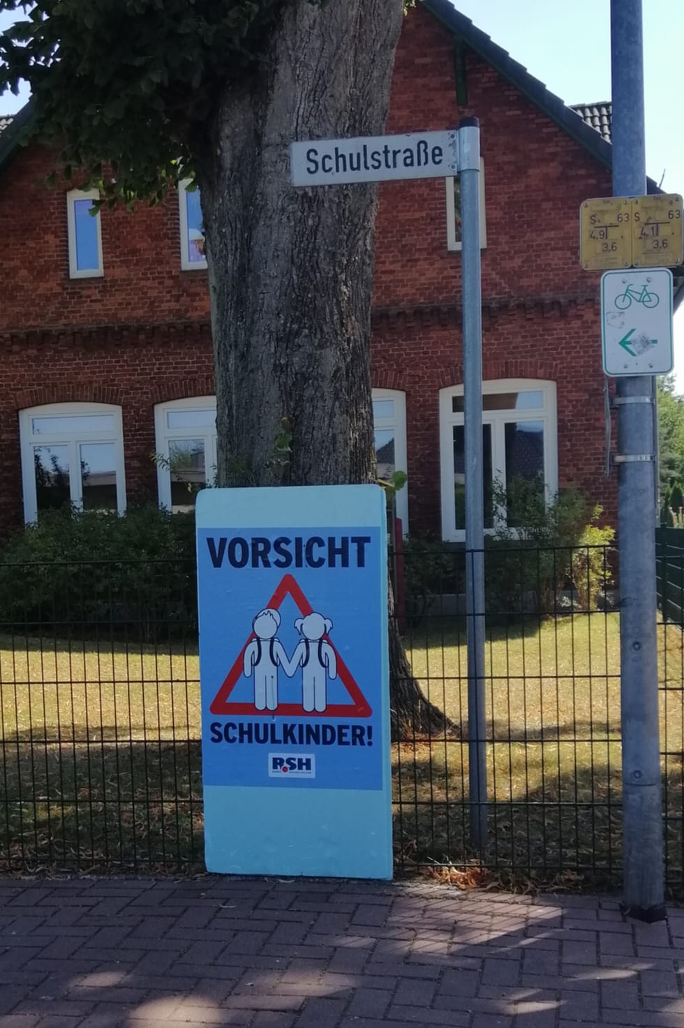 Ein Plakat mit der Aufschrift "Vorsicht Schulkinder" hängt an einem Zaun unter einem Straßenschild mit der Aufschrift "Schulstraße".