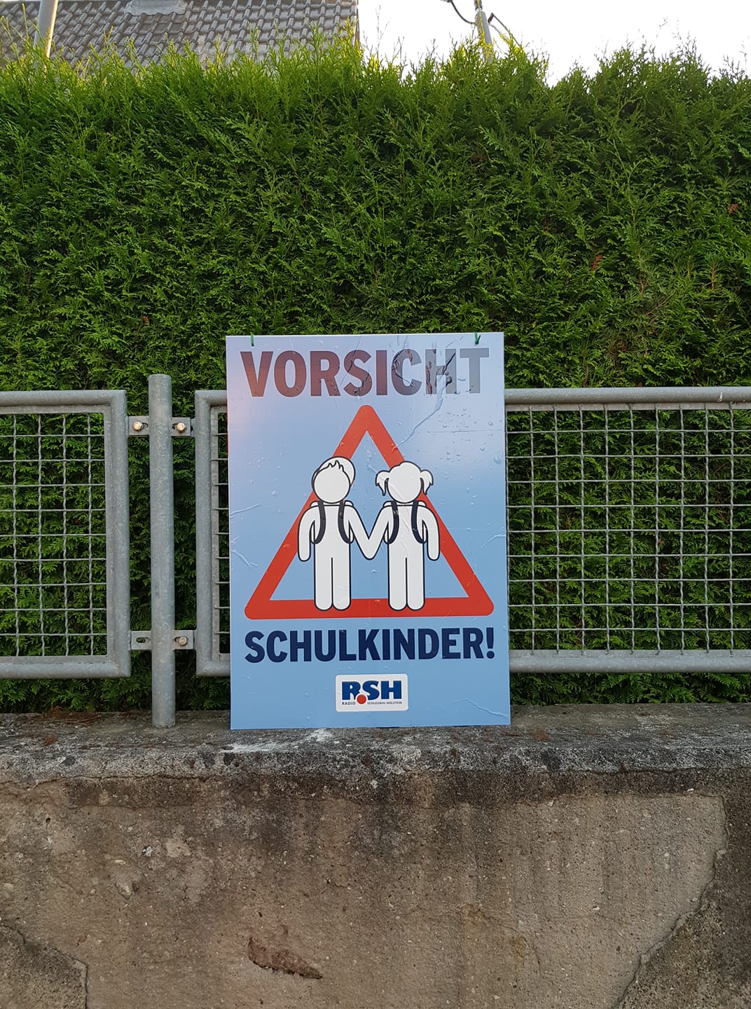 Ein Plakat mit der Aufschrift "Vorsicht Schulkinder" lehnt einem Zaun vor einer Hecke.