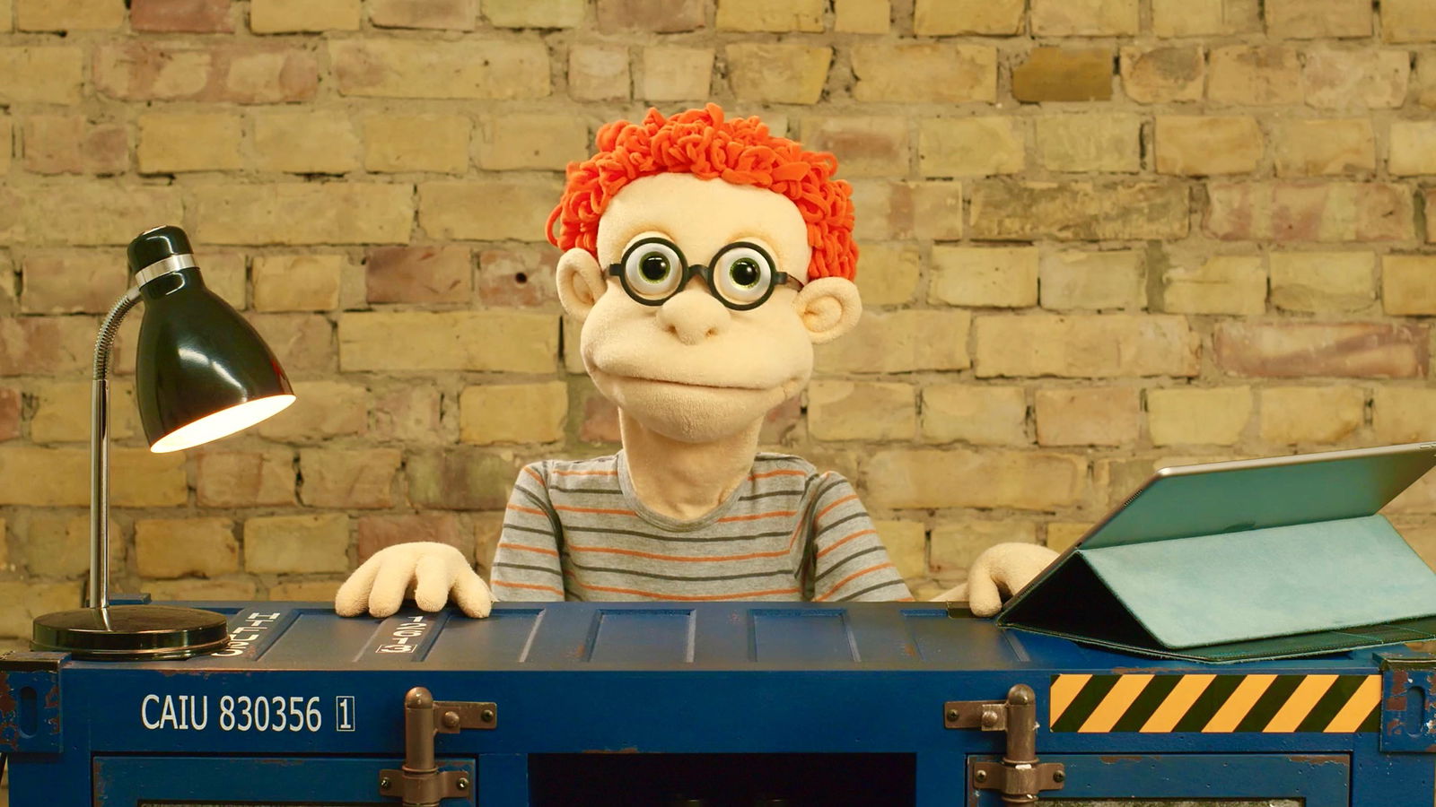 Eine Handpuppe mit orange Haare und Brille sitzt hinter einem dunkelblauen Container. Links auf dem Container steht eine Schreibtischlampe, rechts befindet sich ein Tablet.