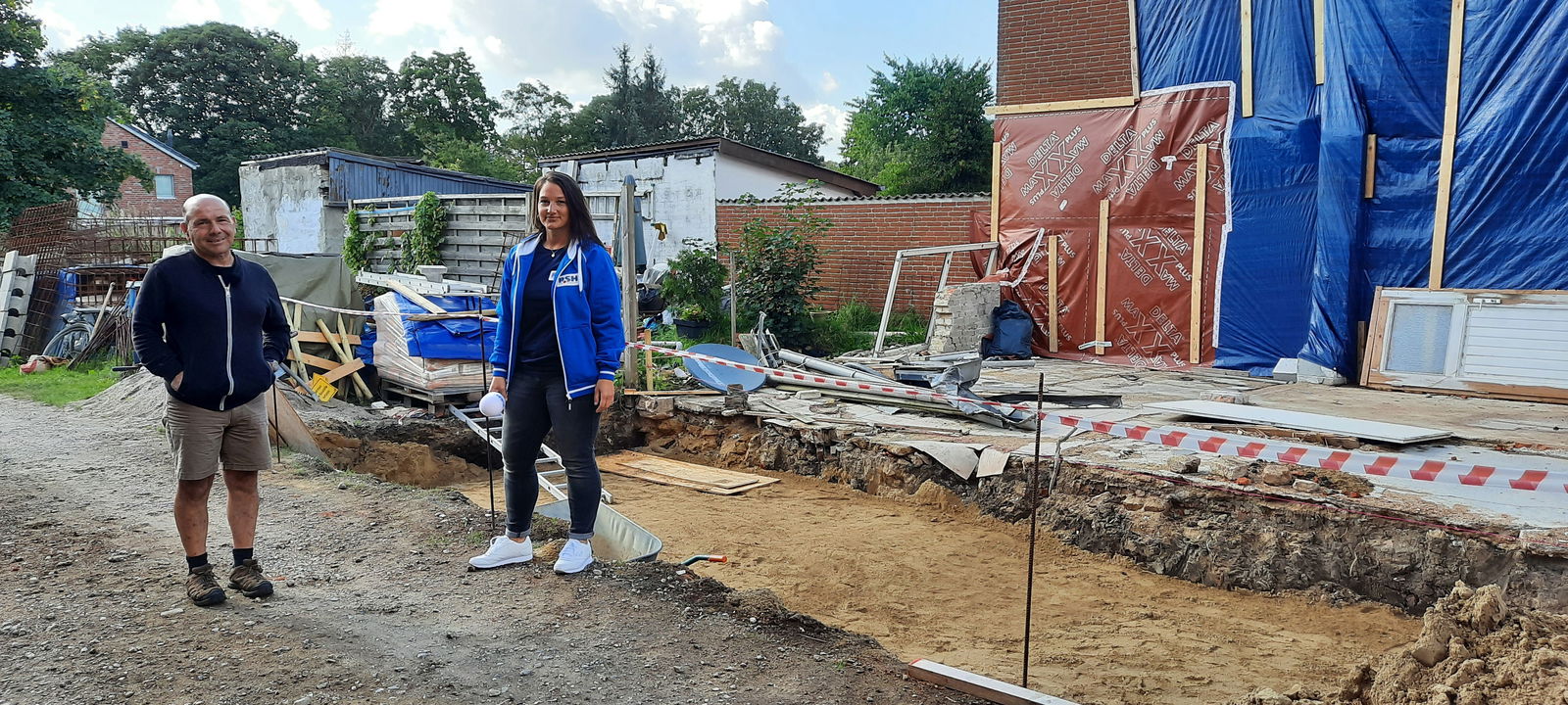 Vor einer Baugrube stehen ein Mann in Jacke und Shorts und eine Frau in R.SH Jacke.