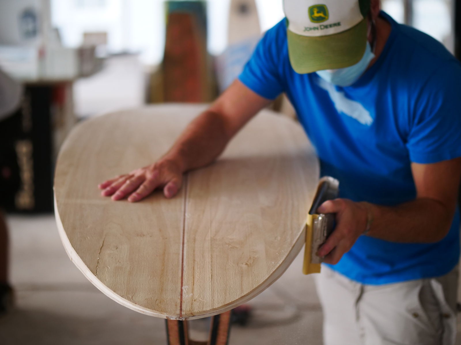 EIn Mann mit Atemschutzmaske steht mit Schleifpapier an einem Surfboard aus Holz und schleift es ab.