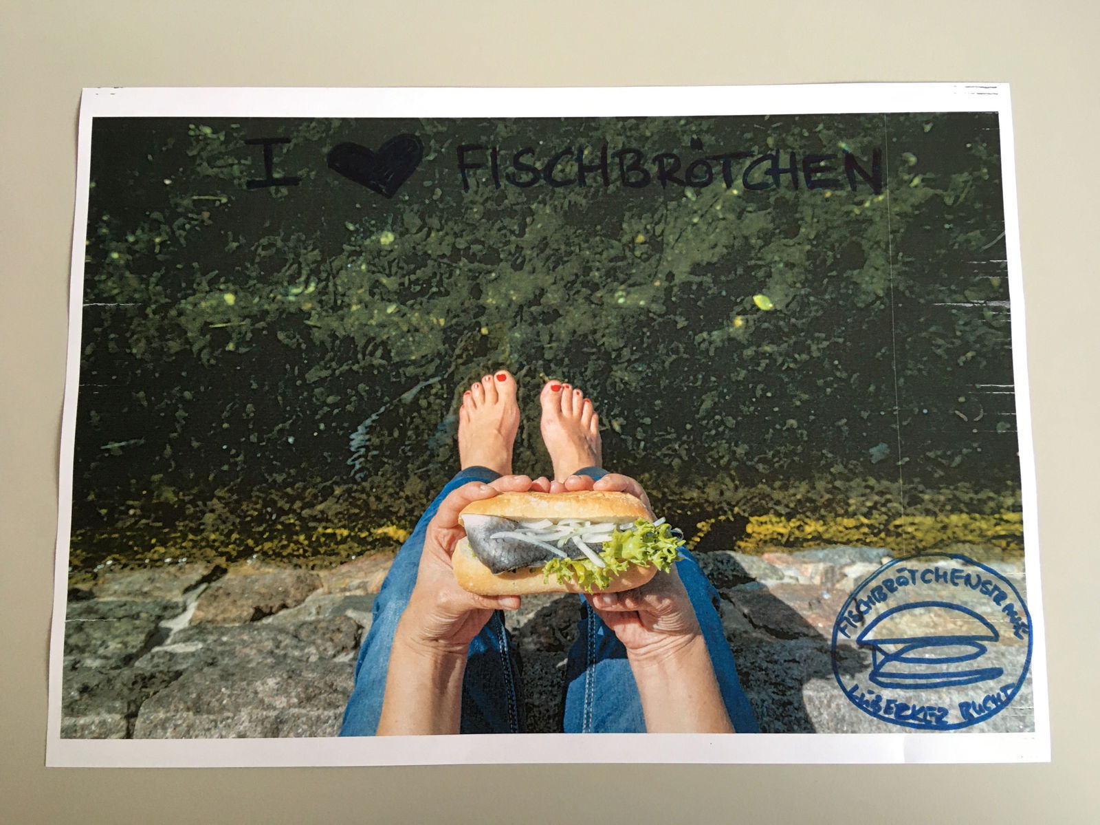 Eine Person sitzt auf einer Kaimauer und hält ein Matjesbrötchen in den Händen in die Kamera. Darüber steht "I love Fischbrötchen."