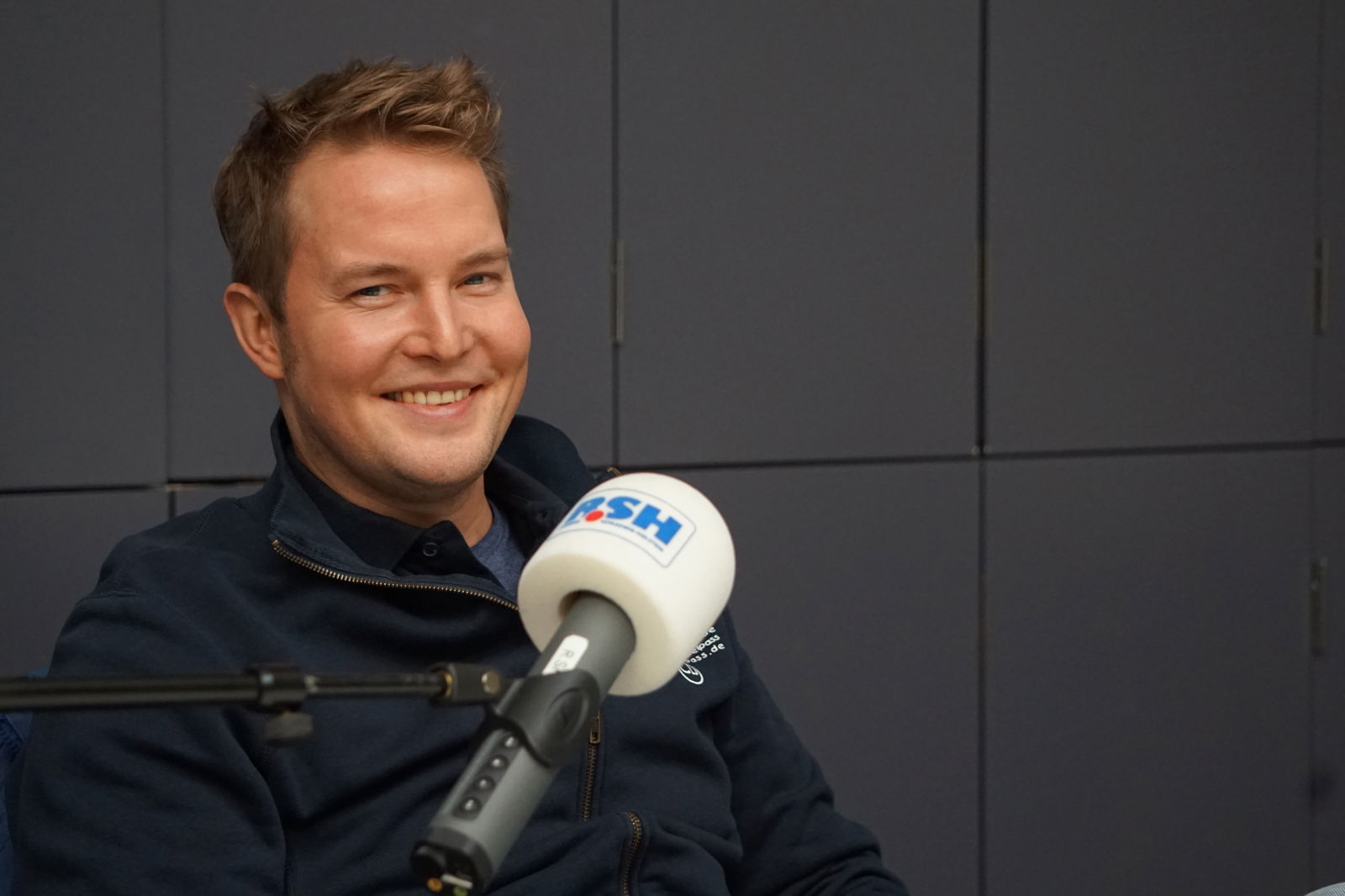 Ein Mann mit kurzen Haaren sitzt auf einem Stuhl und spricht in ein Mikrofon mit R.SH-Aufdruck.