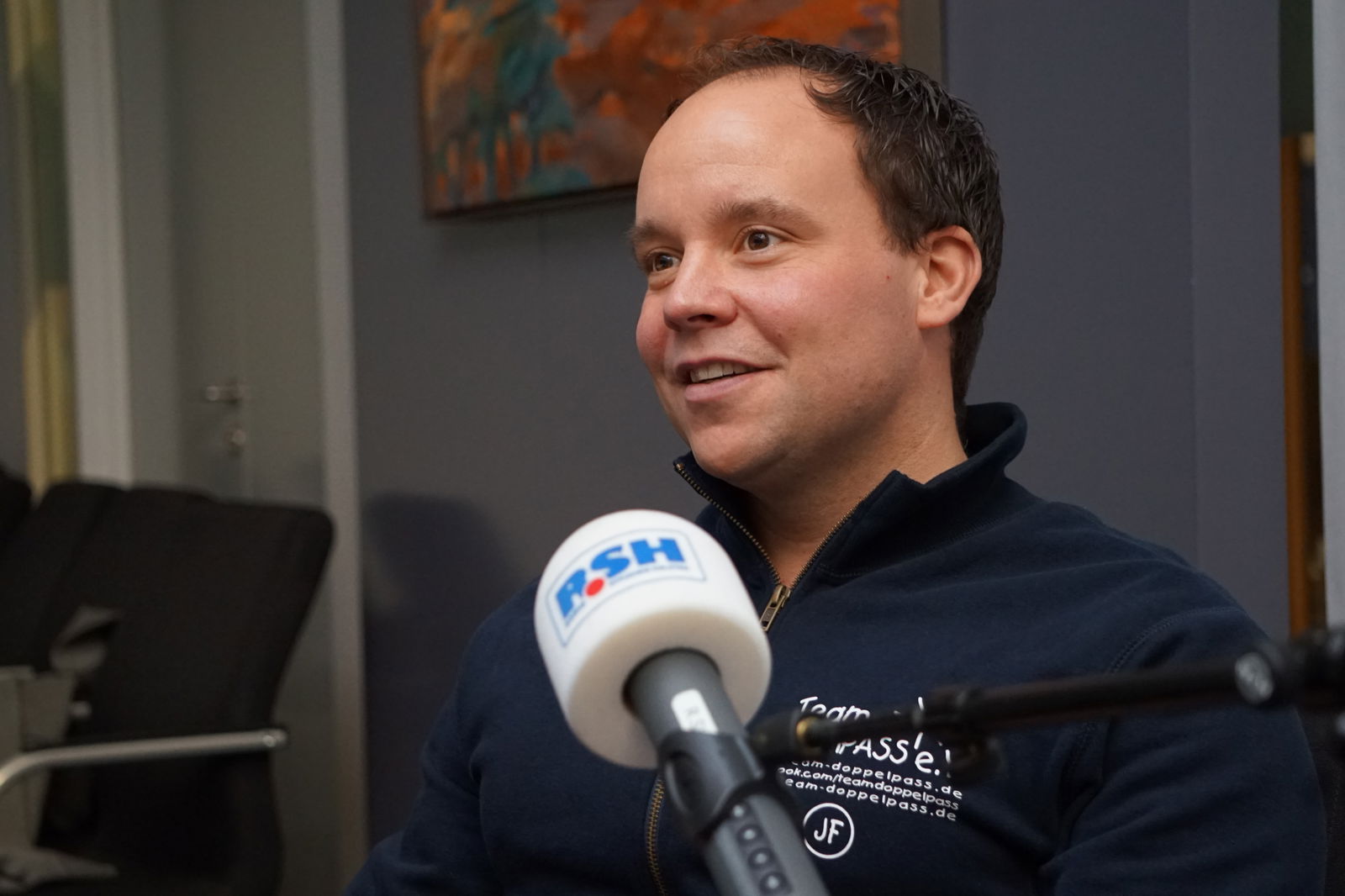 Ein Mann mit kurzen Haaren sitzt auf einem Stuhl und spricht in ein Mikrofon mit R.SH-Aufdruck.