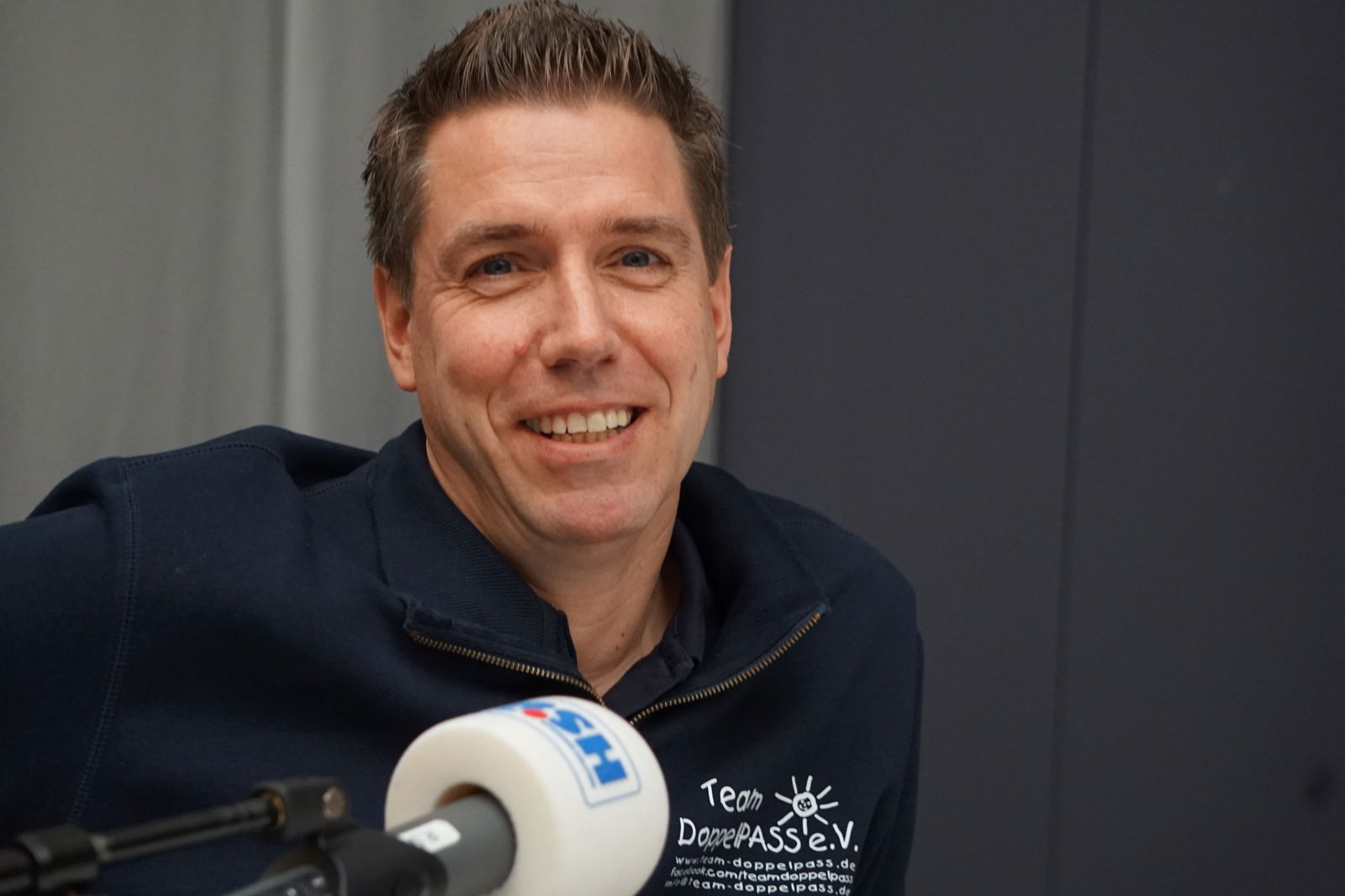 Ein Mann mit kurzen Haaren sitzt auf einem Stuhl und spricht in ein Mikrofon mit R.SH-Aufdruck.