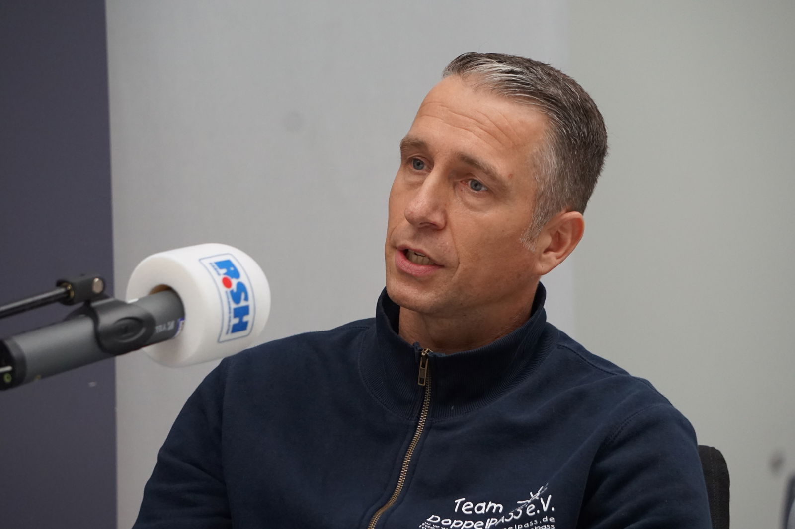 Ein Mann mit kurzen Haaren sitzt auf einem Stuhl und spricht in ein Mikrofon mit R.SH-Aufdruck.