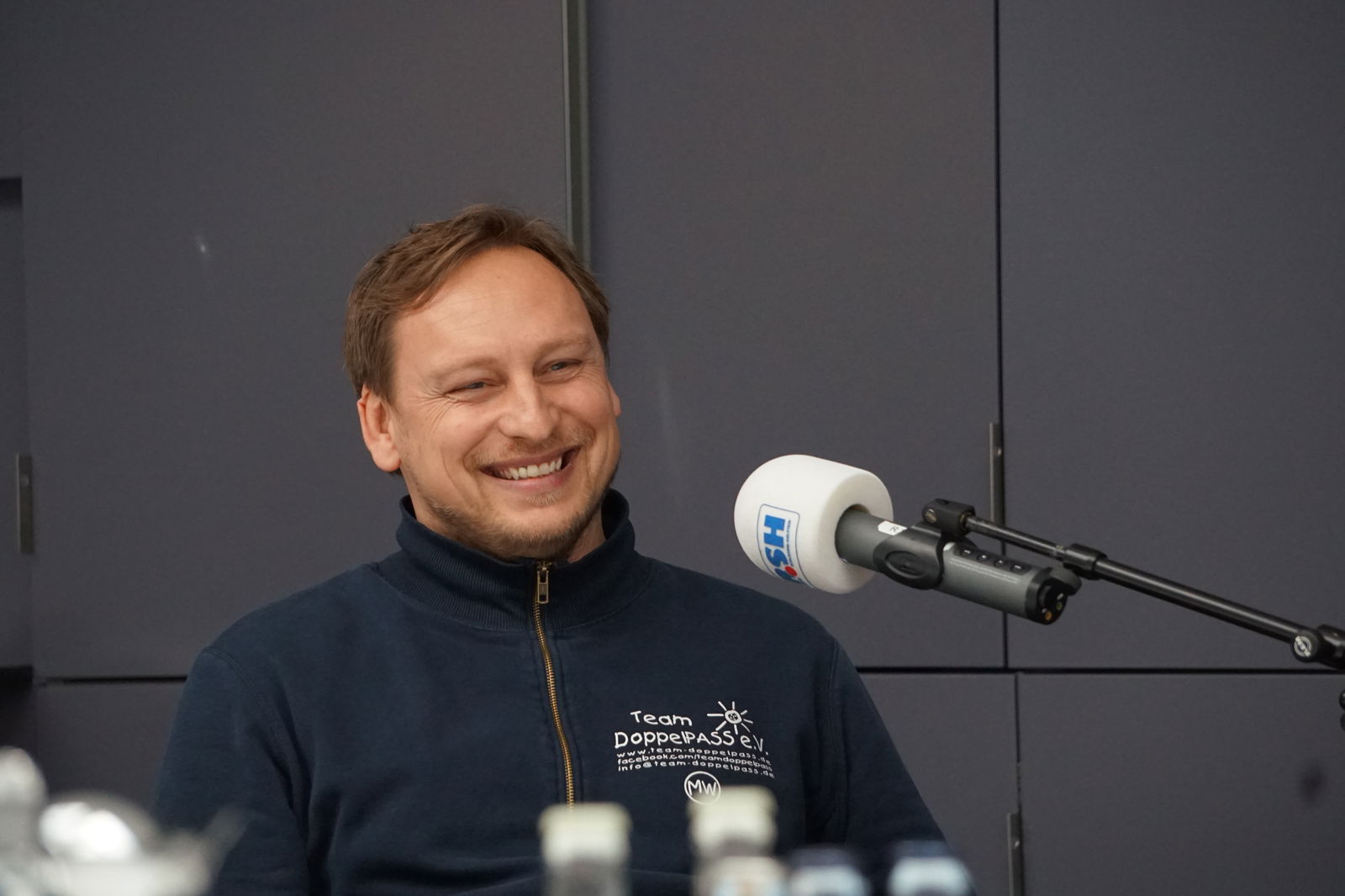 Ein Mann mit kurzen Haaren sitzt auf einem Stuhl und spricht in ein Mikrofon mit R.SH-Aufdruck.